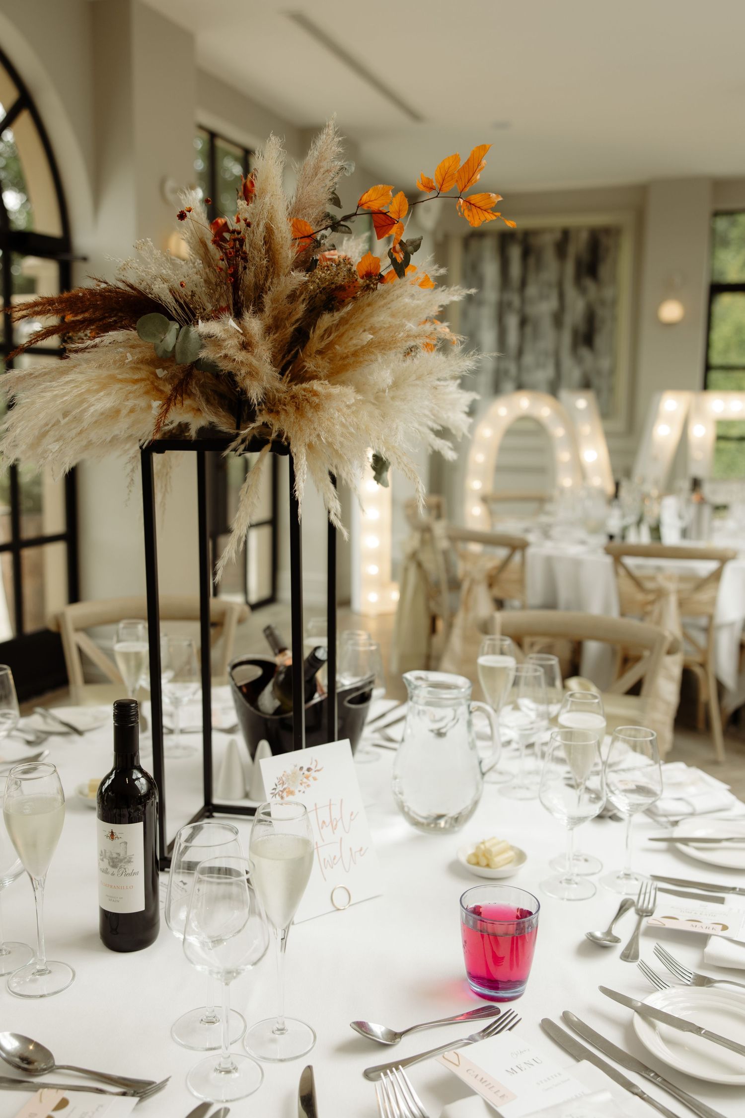 Elegant wedding reception tables with white linens feature tall feather centerpieces and formal place settings.