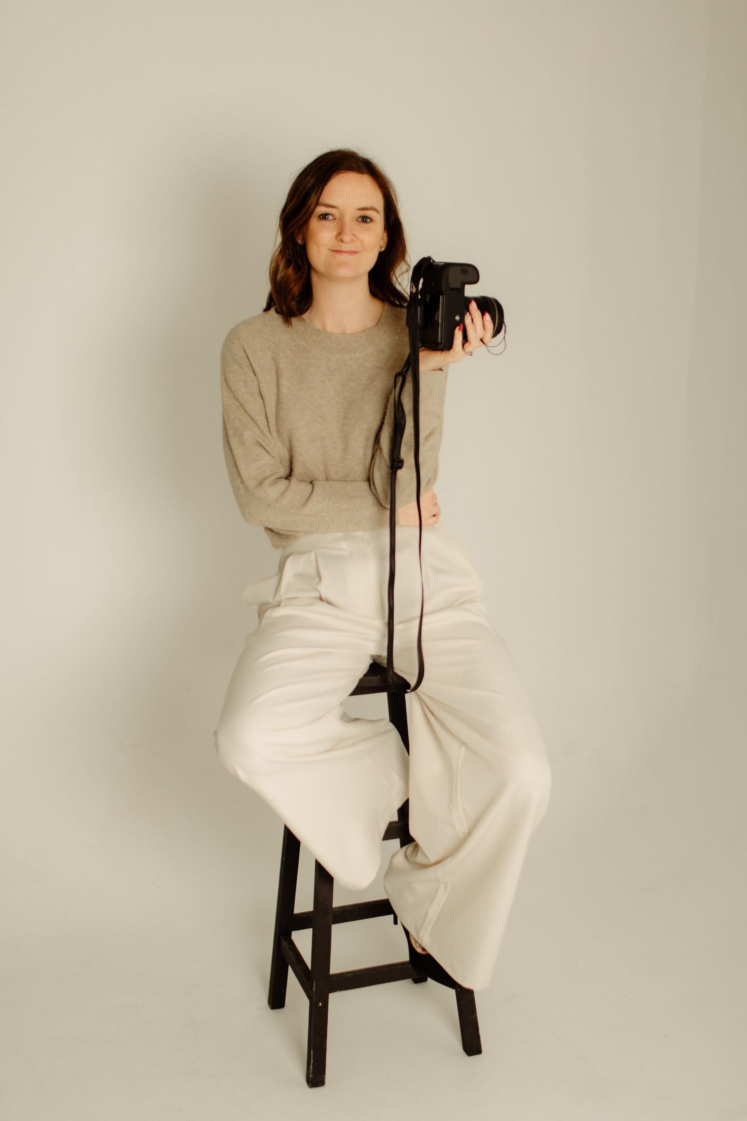 Photographer in beige sweater and white pants sits on dark stool with professional camera against neutral background.
