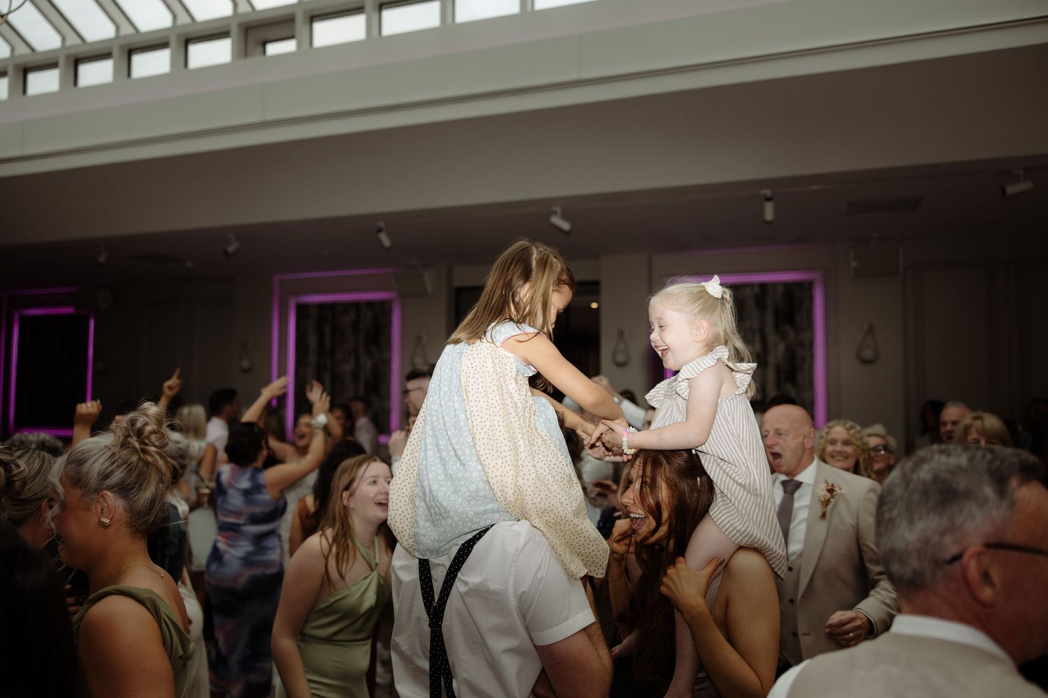 A joyful crowd dances and celebrates at a wedding reception with purple accent lighting.