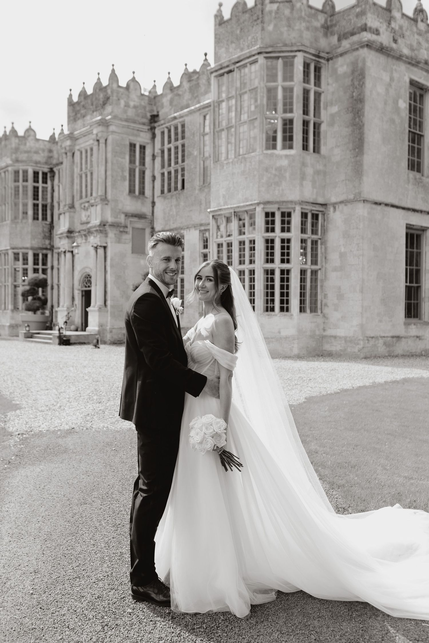 Elegant wedding couple poses in front of a grand historic manor house with flowing white dress and classic architecture.