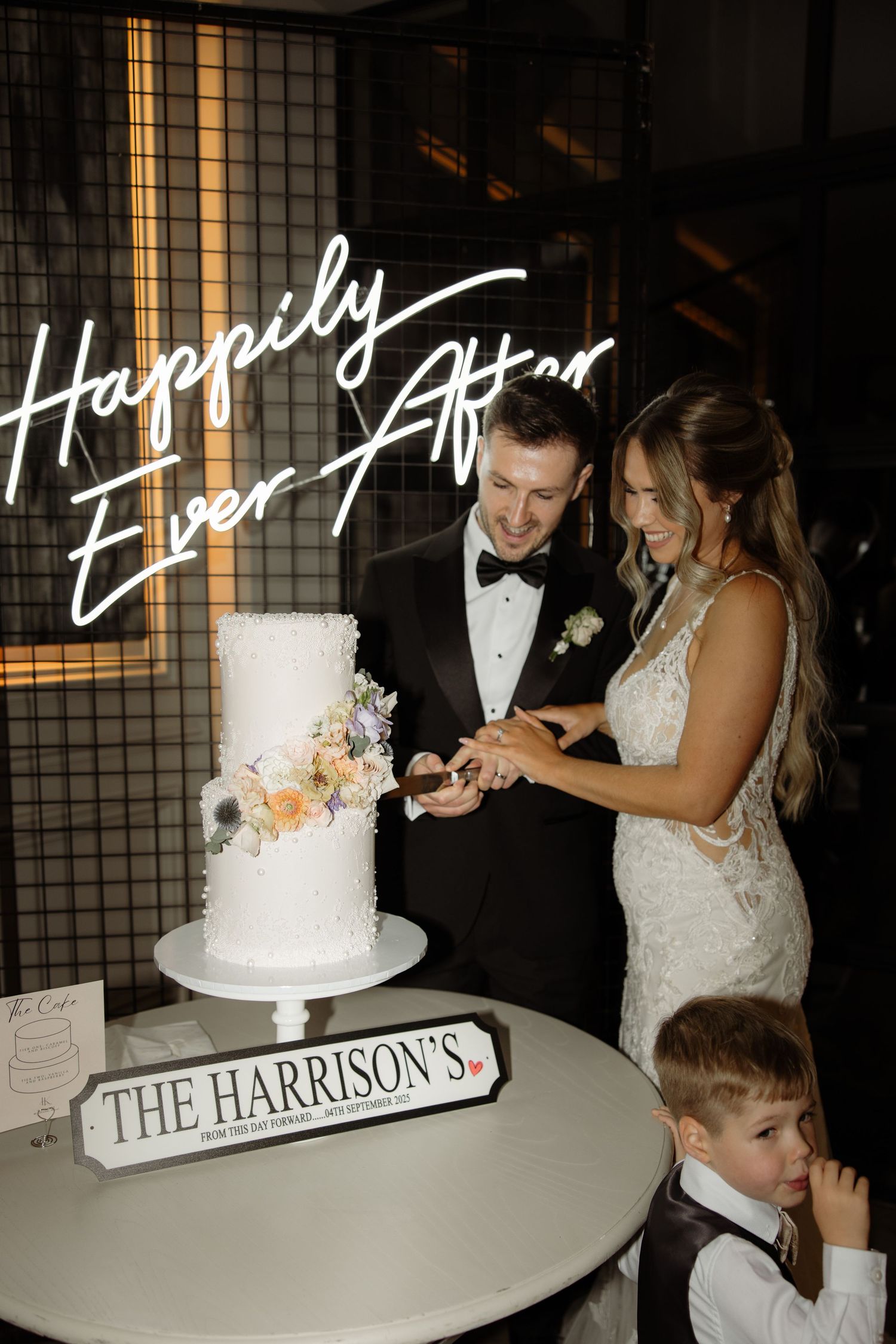 Newlyweds share a joyful moment cutting their elegant white wedding cake next to a 'Happily Ever After' sign.