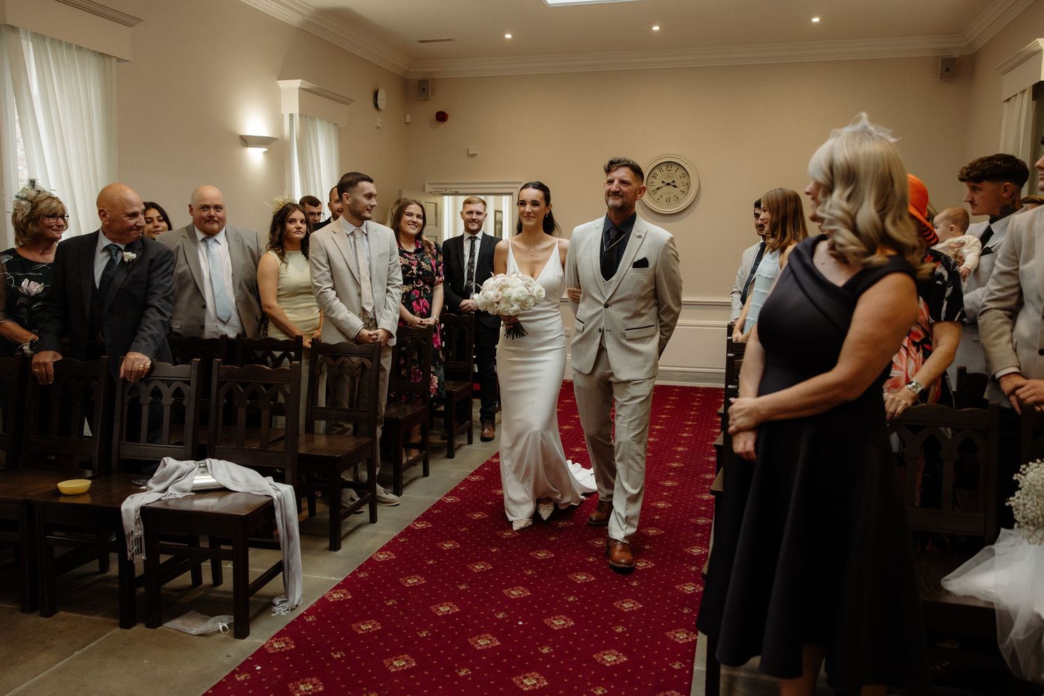 Wedding ceremony guests stand in a bright indoor venue with red carpeting and elegant decor.