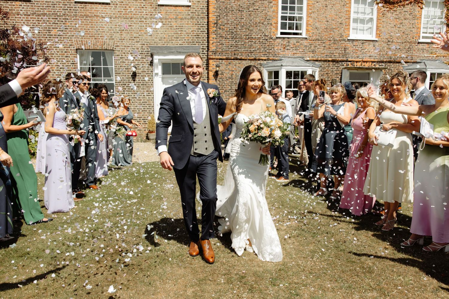 Wedding guests shower the newlyweds with white flower petals as they walk together outside a brick building.