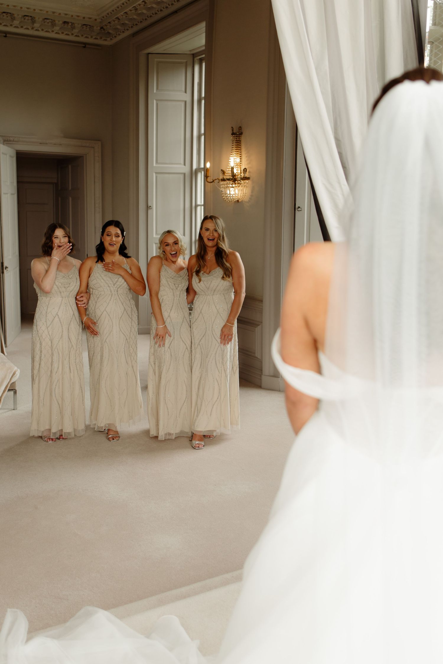A group in matching cream-colored dresses poses together in an elegant room with ornate moldings and white curtains.