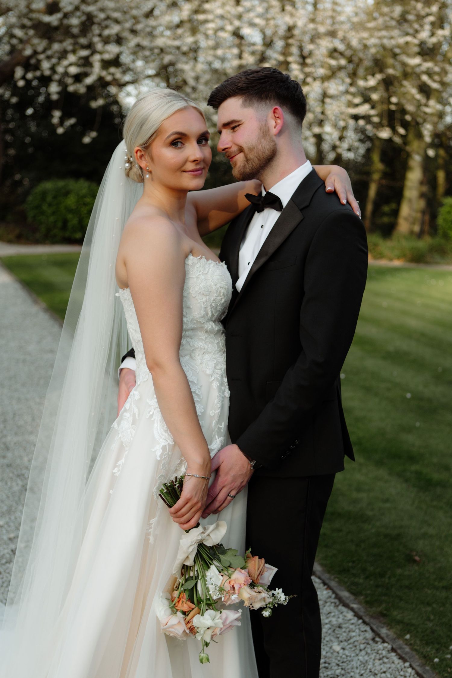 A romantic wedding couple poses together outdoors with blooming trees and greenery in the background.