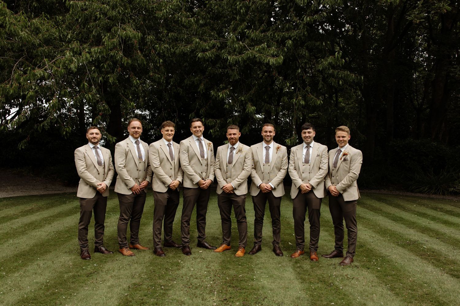 Wedding party in khaki and brown outfits poses together on a lush green lawn during an outdoor celebration.