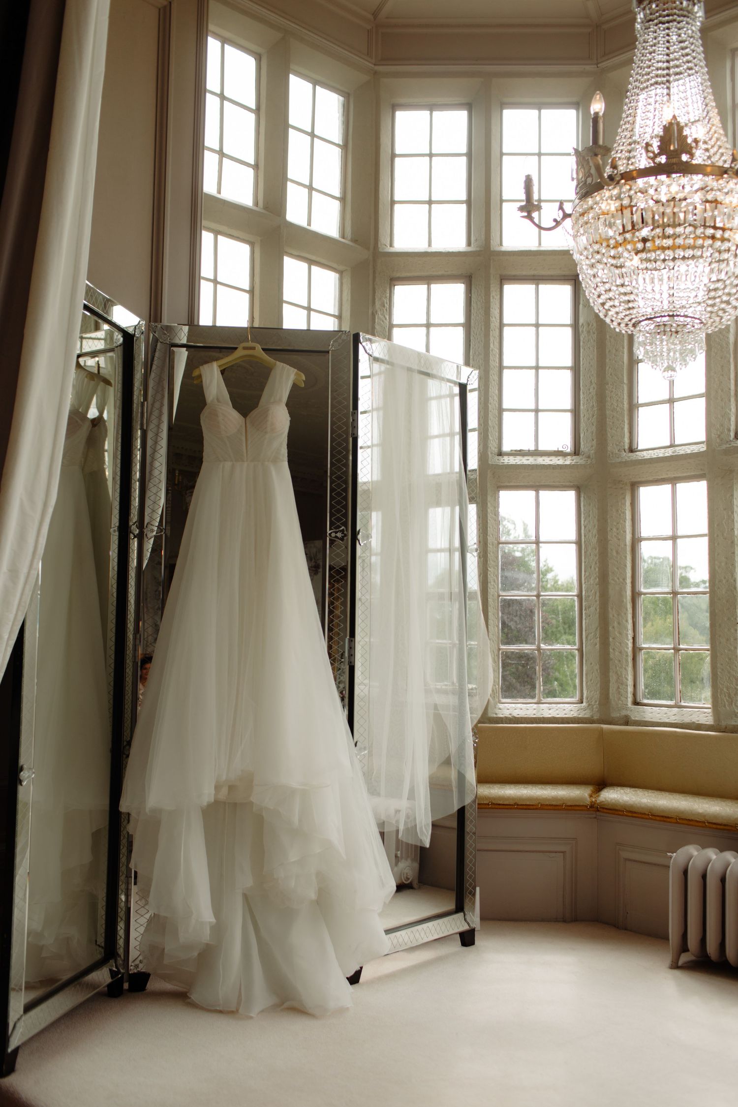 Multiple white wedding gowns hanging in an elegant room with crystal chandeliers and large bay windows.