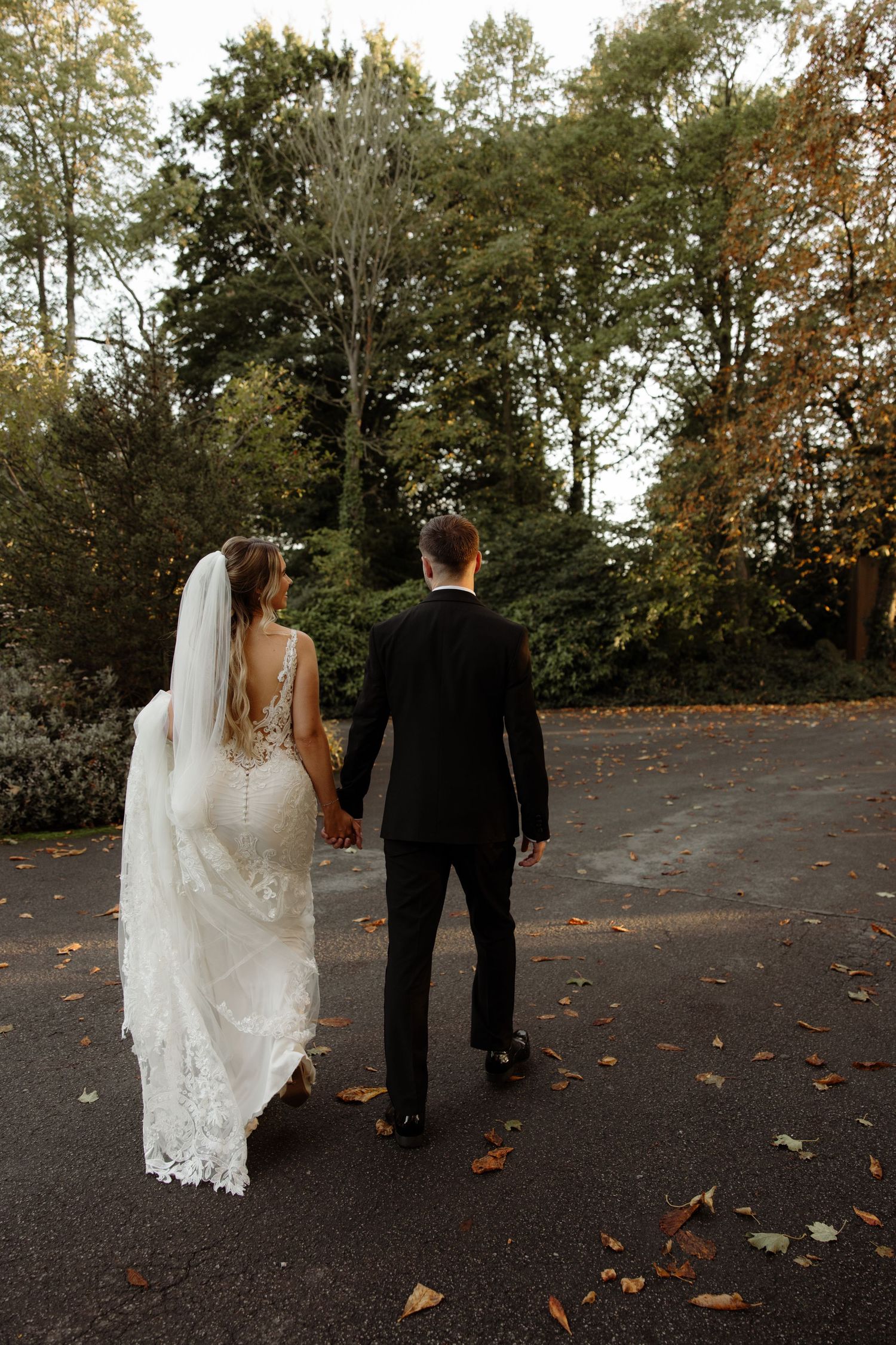 A couple in wedding attire walking away on a path with autumn leaves scattered on the ground.
