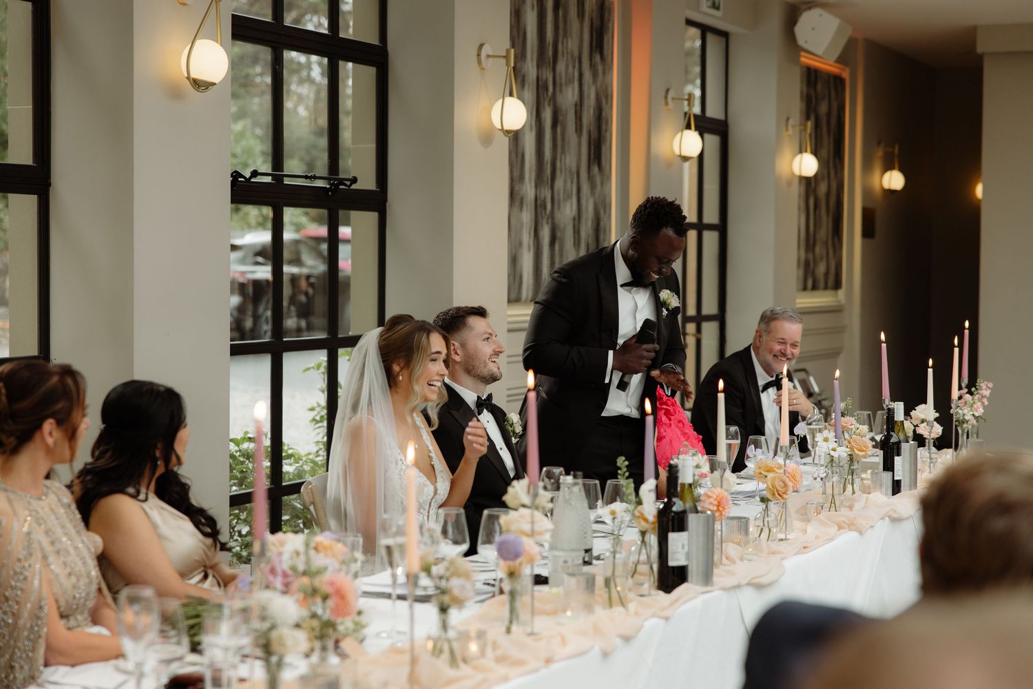Wedding guests seated at an elegant banquet table with floral centerpieces and glassware in a bright reception hall.