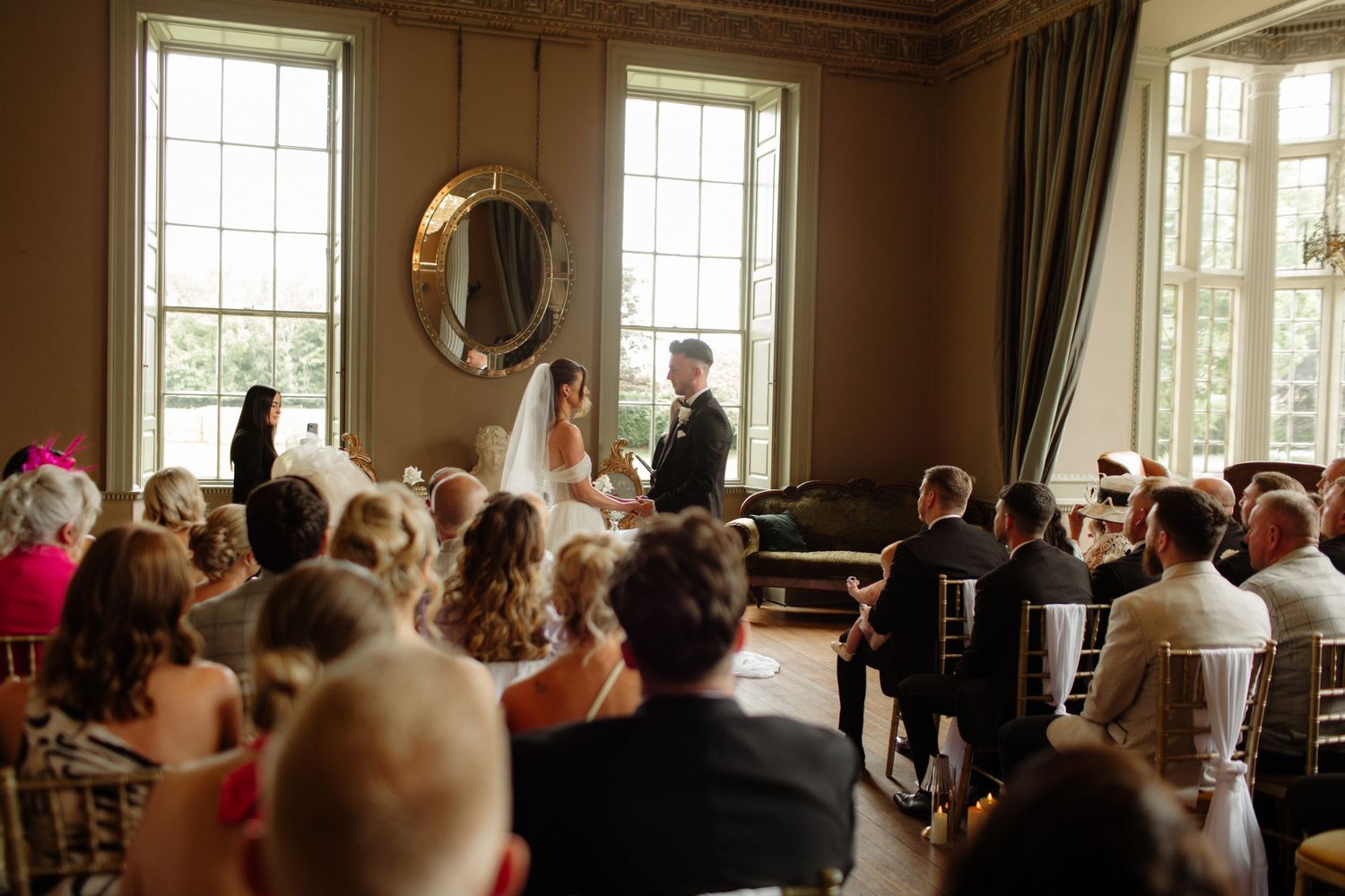 A gathering of people seated in an elegant room with tall windows and a round mirror during what appears to be a ceremony.