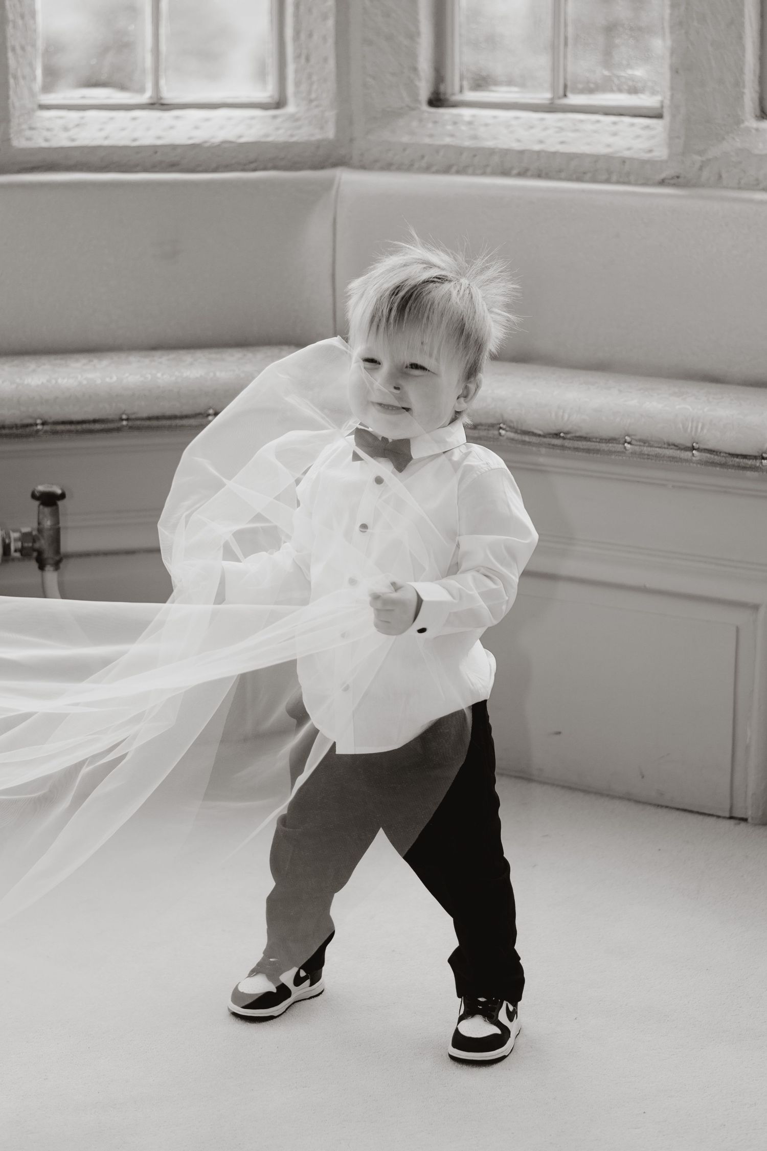 A small child in formal wear plays with flowing white fabric in a bright room.