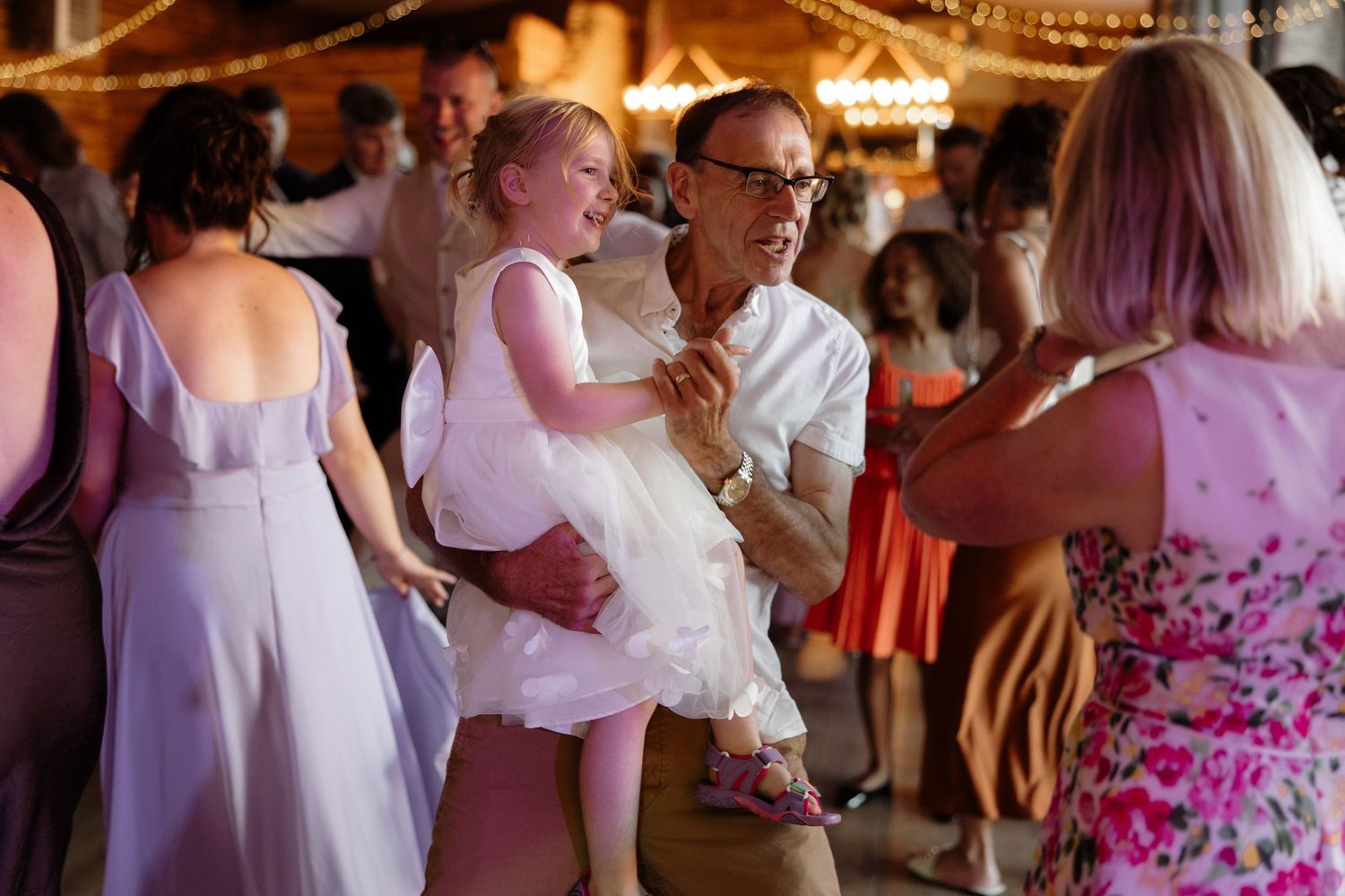 Wedding guests dancing and celebrating together on a rustic venue dance floor with string lights overhead.