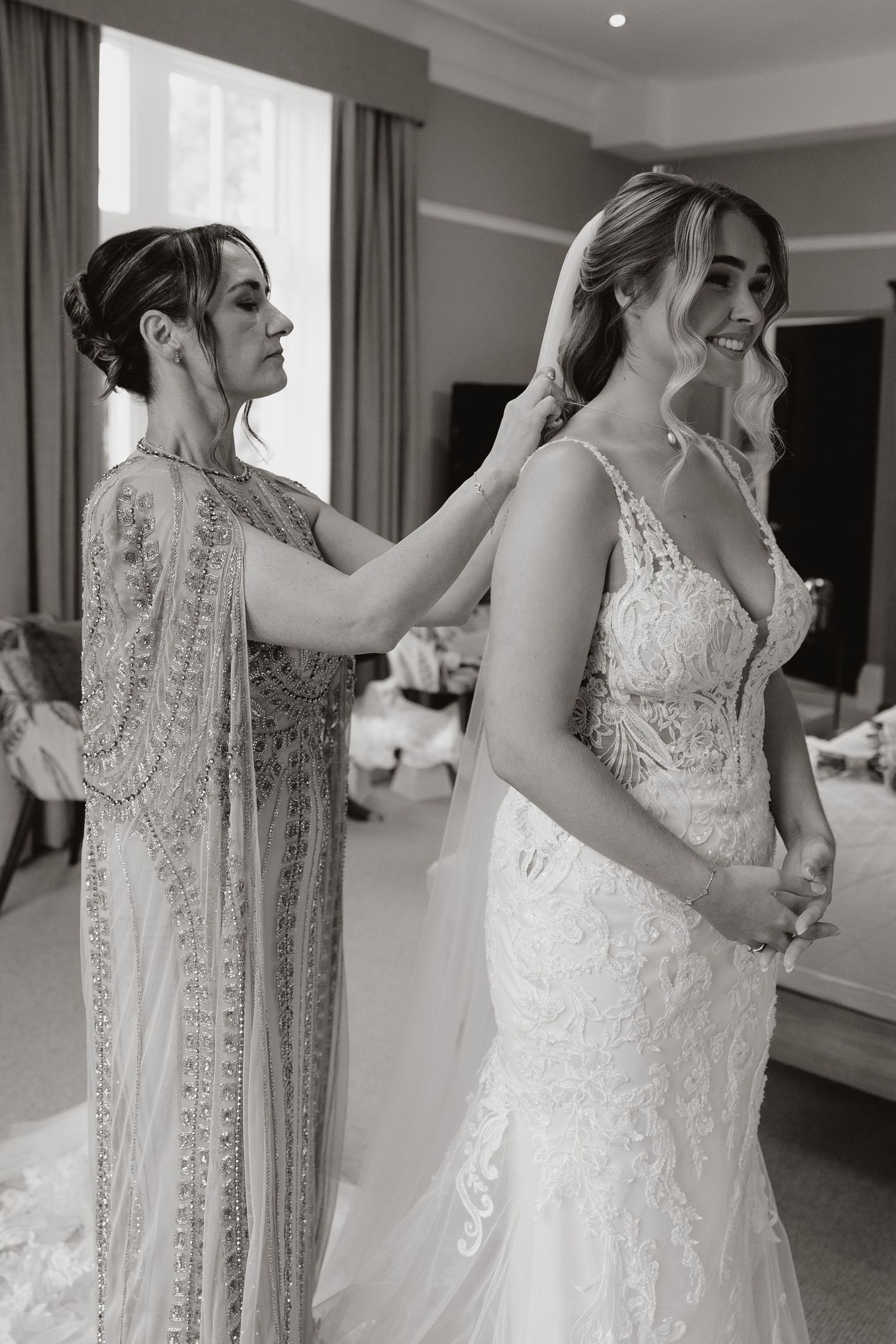 A bride in a detailed lace wedding gown gets ready in a bright room with elegant curtains and decor.