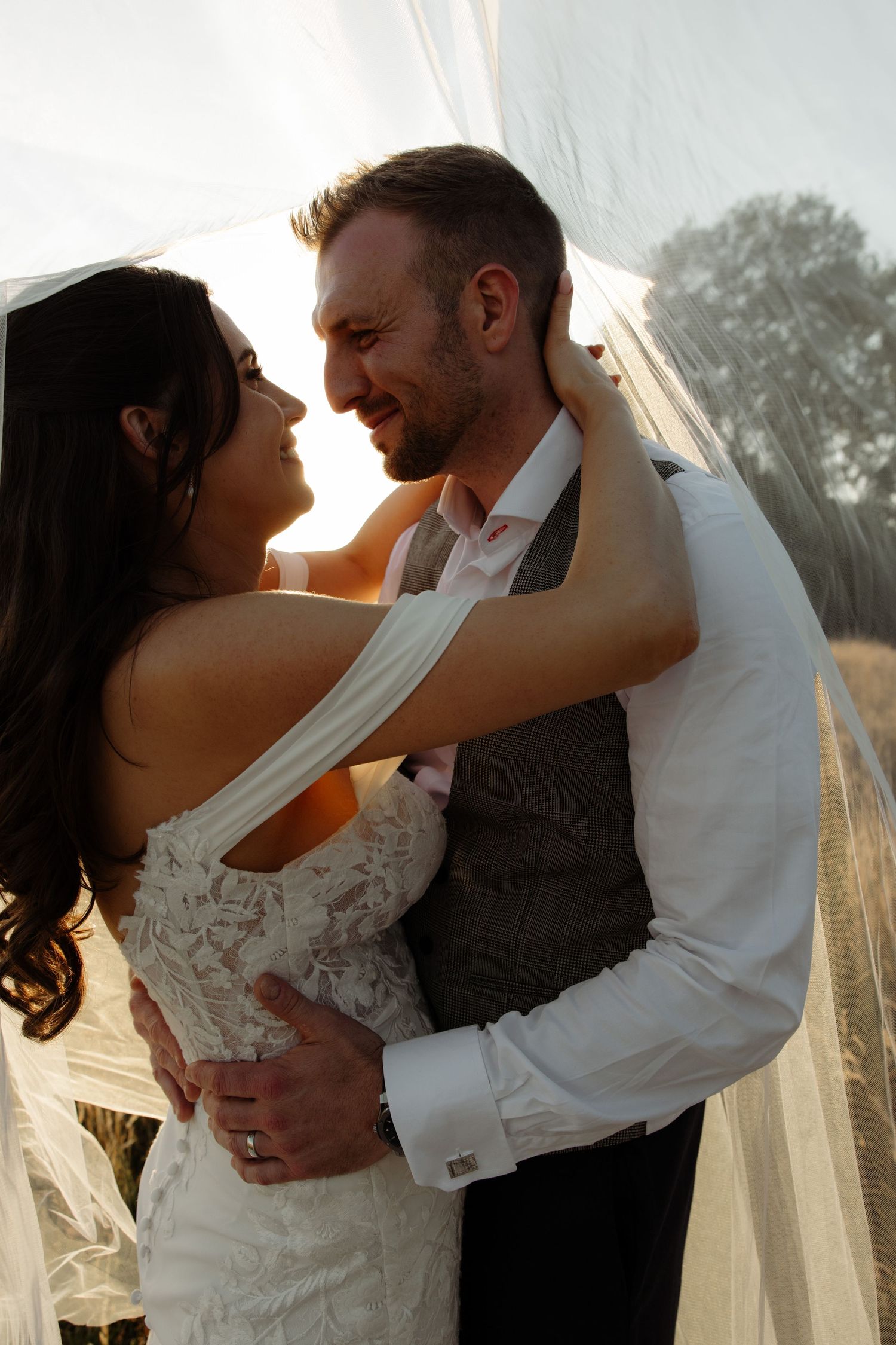 A bride and groom share an intimate moment at sunset, with soft light filtering through her veil.