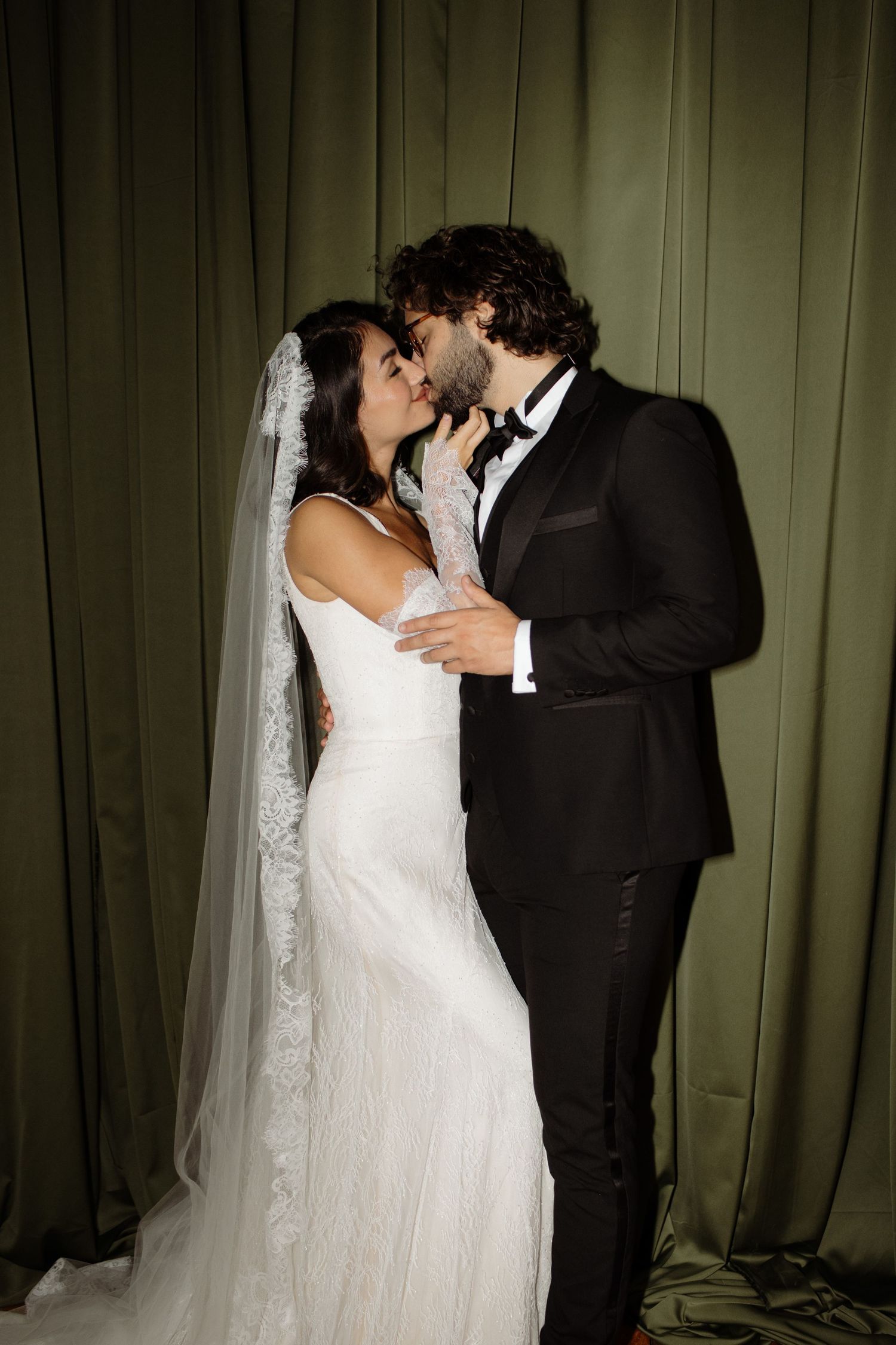 A bride in a white wedding gown and groom in black tuxedo share an intimate moment against a green curtain backdrop.