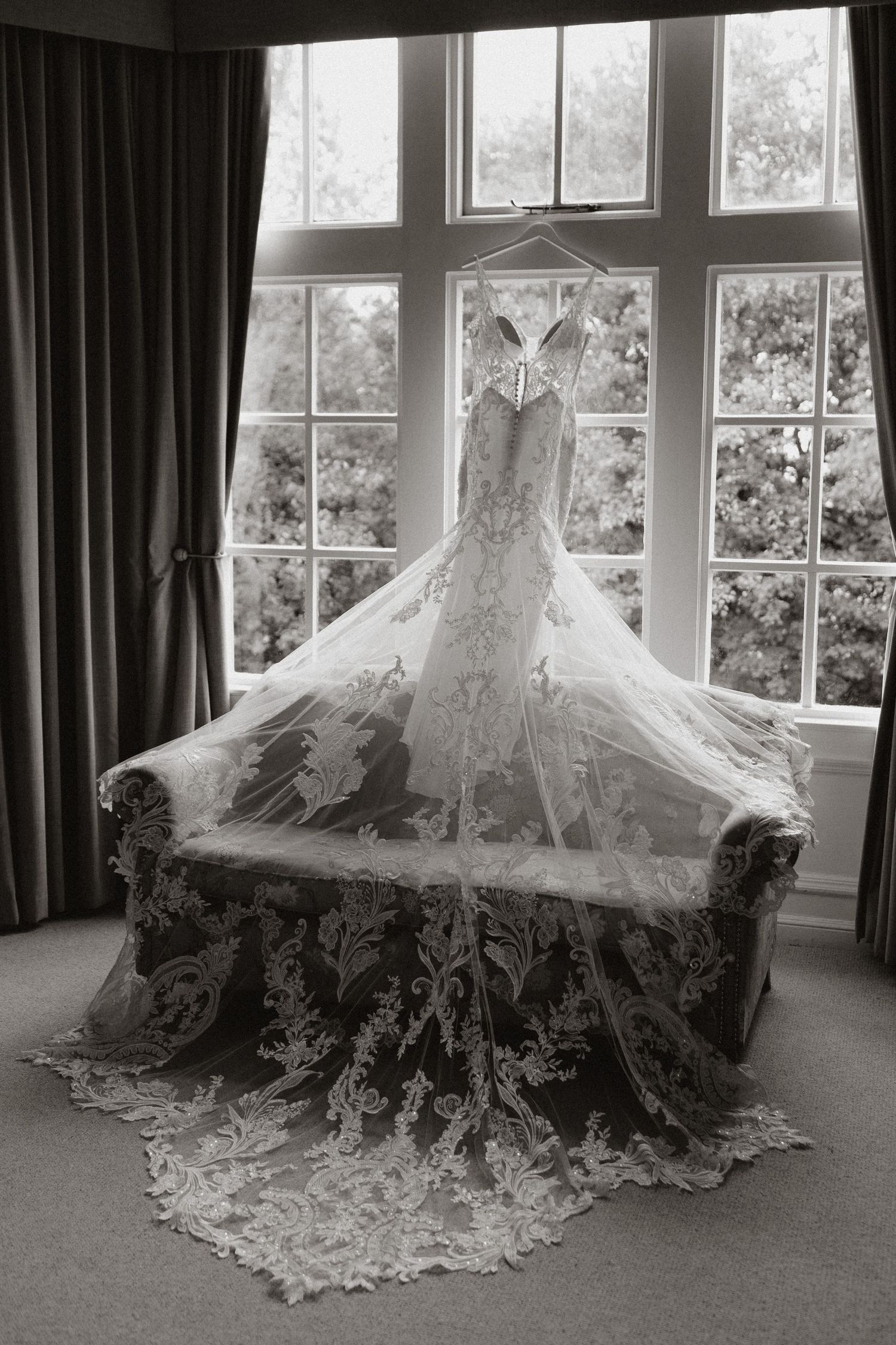 Elegant white wedding dress with intricate lace detailing displayed in a sunlit room with multiple window views.