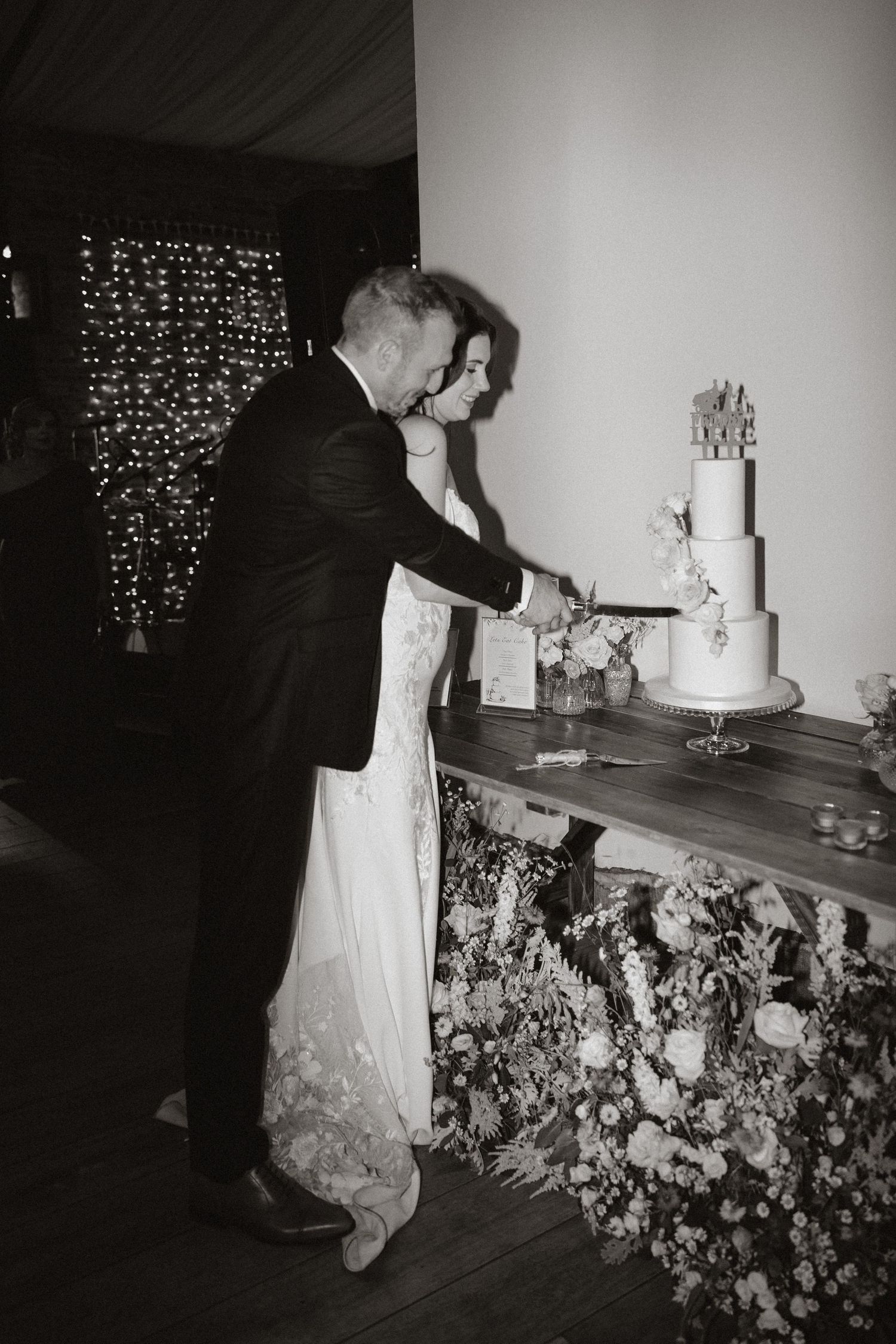 A couple cuts their elegant white wedding cake together at their reception with twinkle lights in the background.