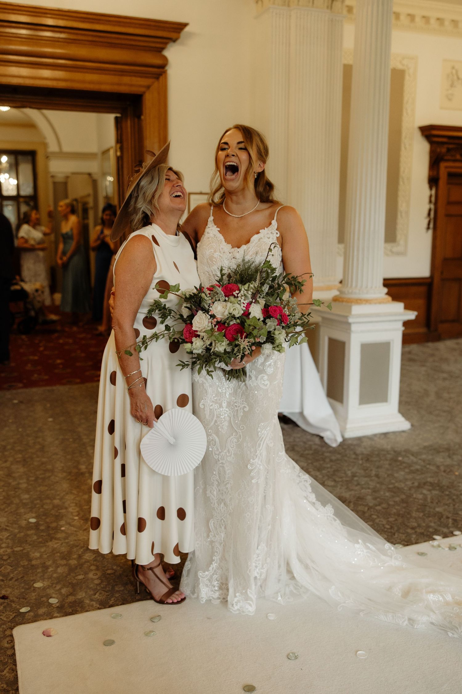Joyful wedding moment captured in an elegant venue with white columns and floral bouquets featuring roses and greenery.