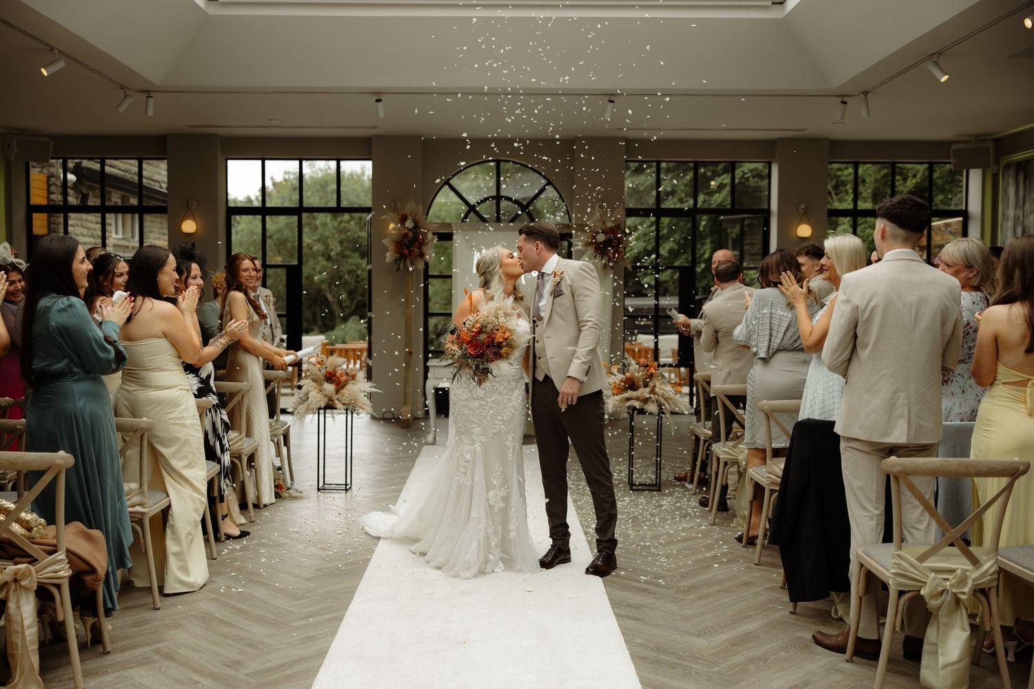 Elegant indoor wedding ceremony with guests standing in a bright conservatory space decorated with floral archway.