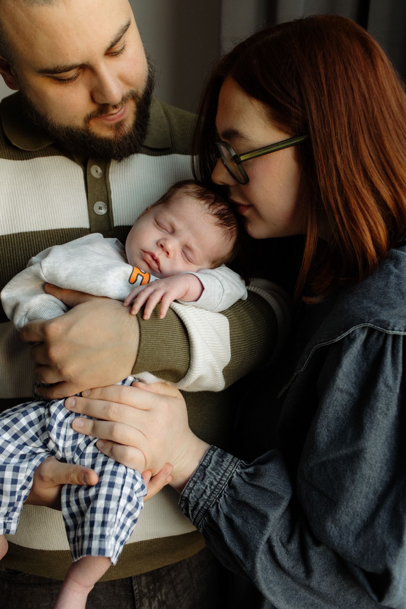 New parents lovingly hold their sleeping newborn baby, mother kissing infant's head gently.