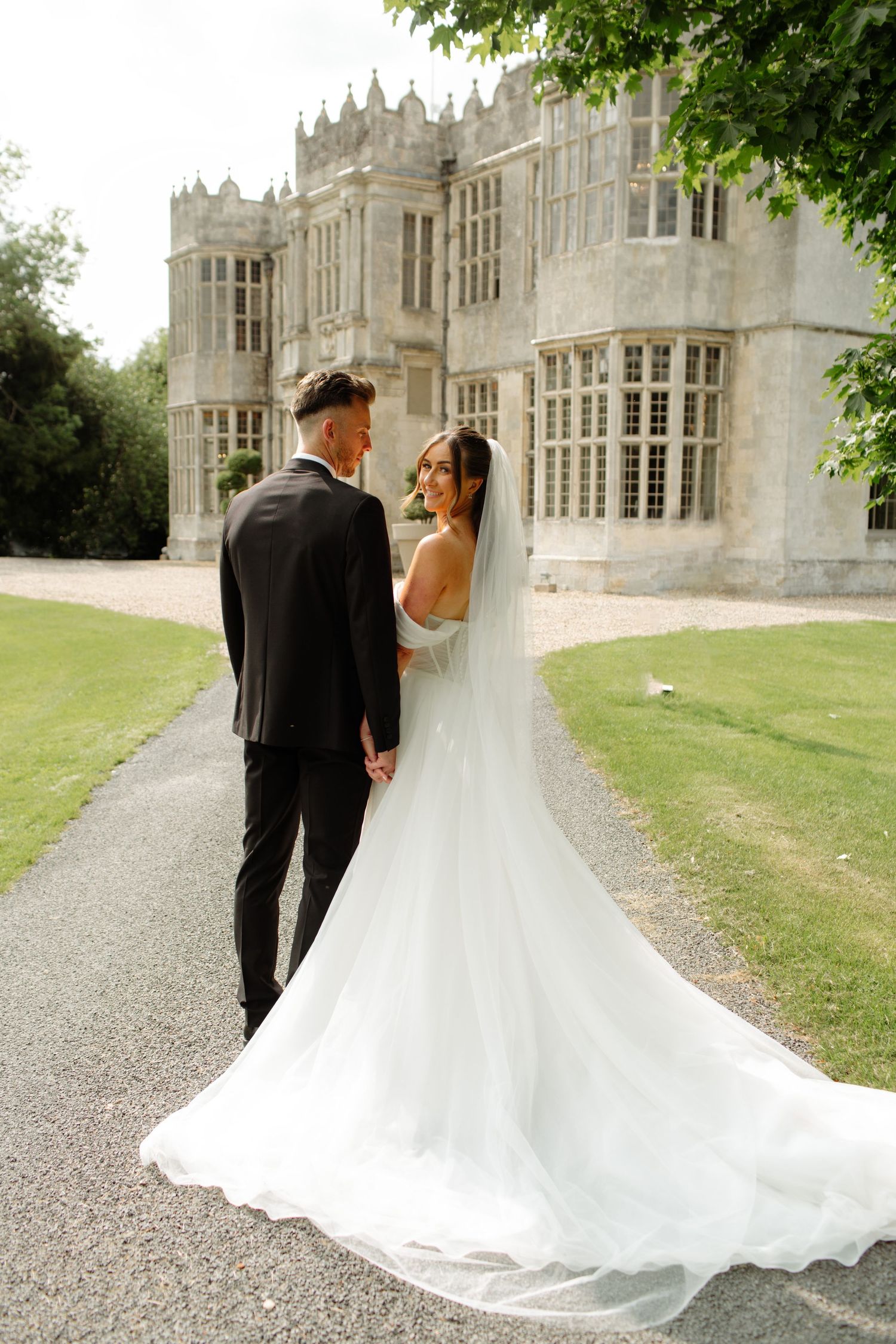A couple in wedding attire walks along a gravel path beside a grand stone Gothic castle-style building.
