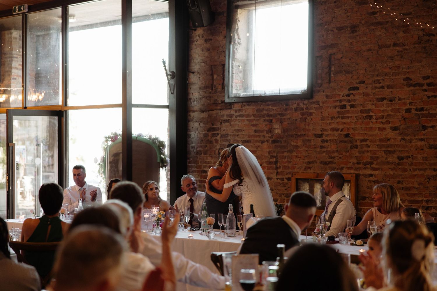 Wedding guests raise their glasses in a toast during a celebration in a rustic brick-walled venue with string lights.