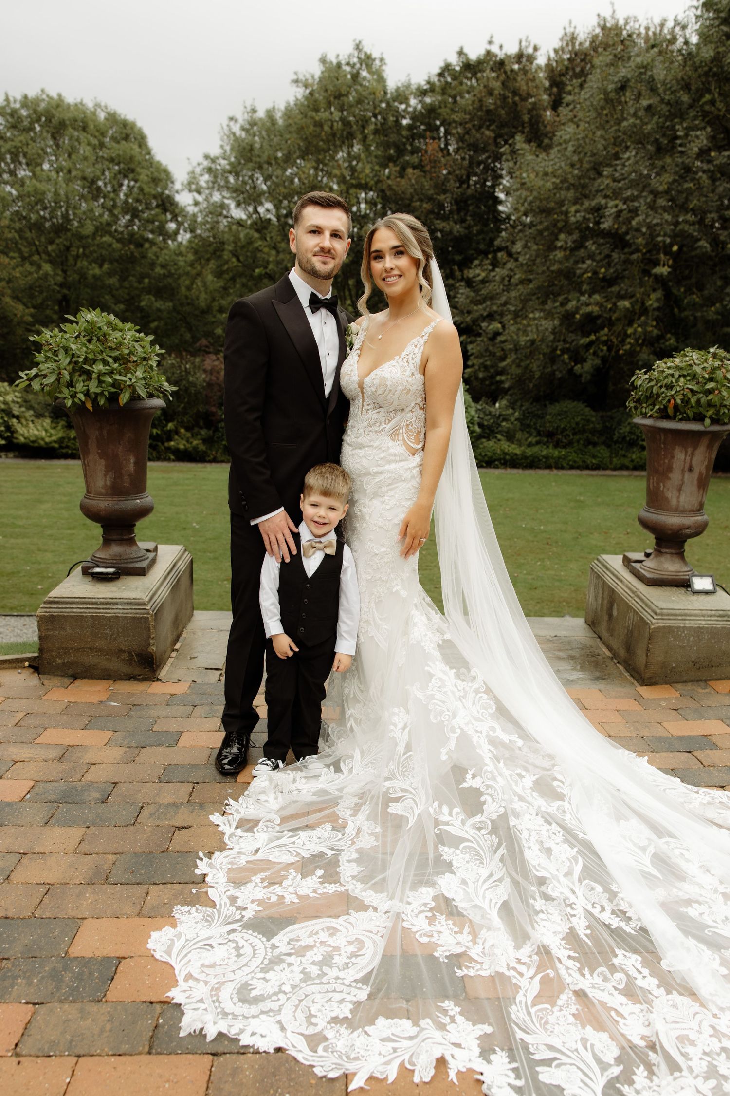 A wedding couple poses with their young child on a stone patio surrounded by lush greenery.