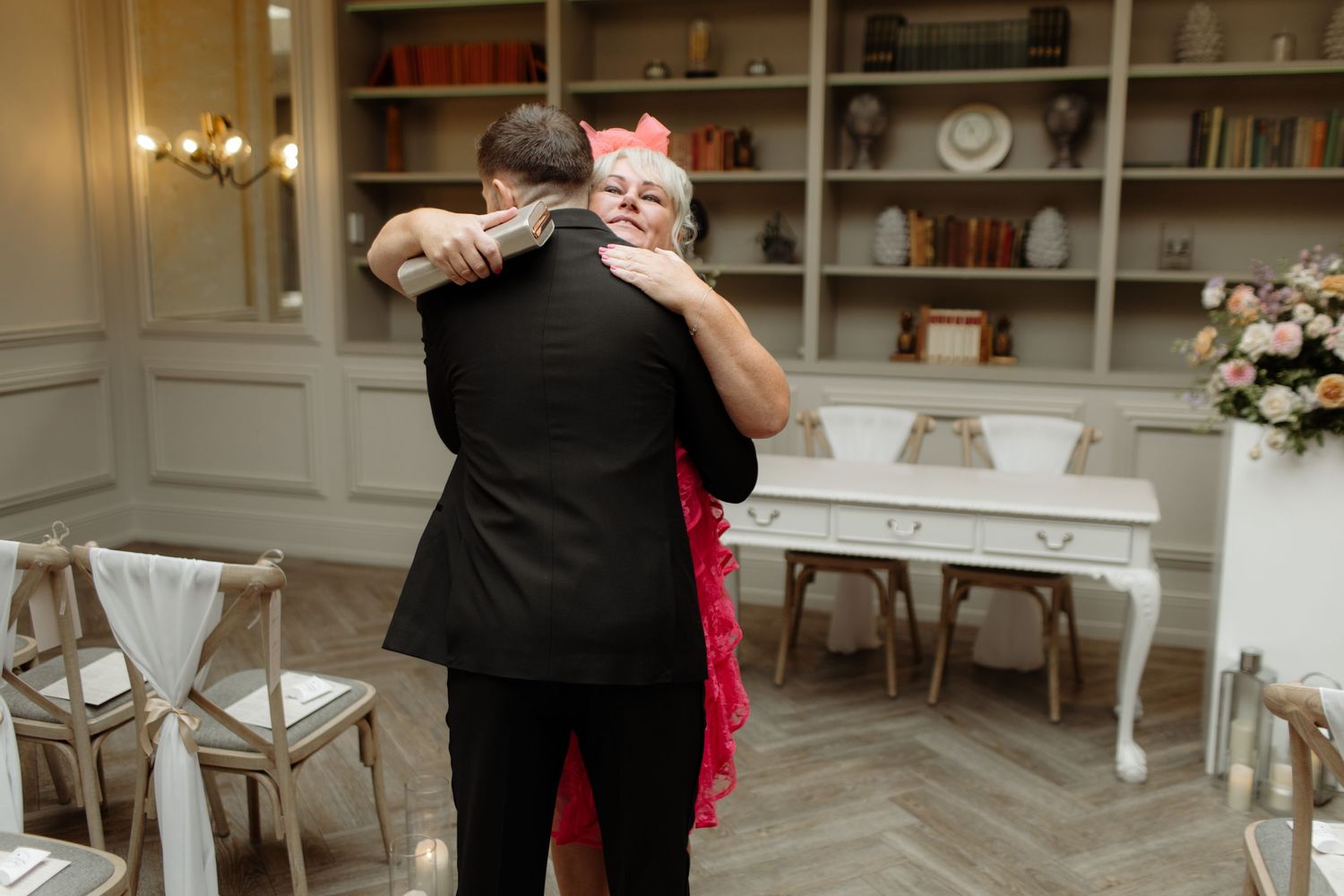 Two people share an intimate dance embrace in an elegant room with white chairs and built-in bookshelves.