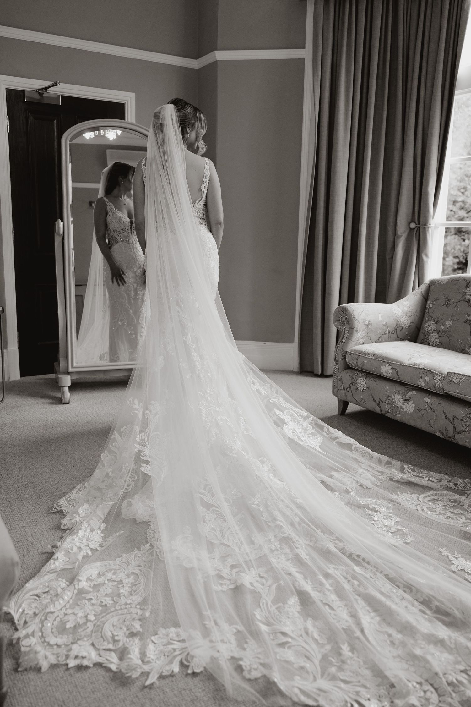 A stunning lace wedding dress with a long cathedral train is reflected in an elegant mirror beside a vintage sofa.