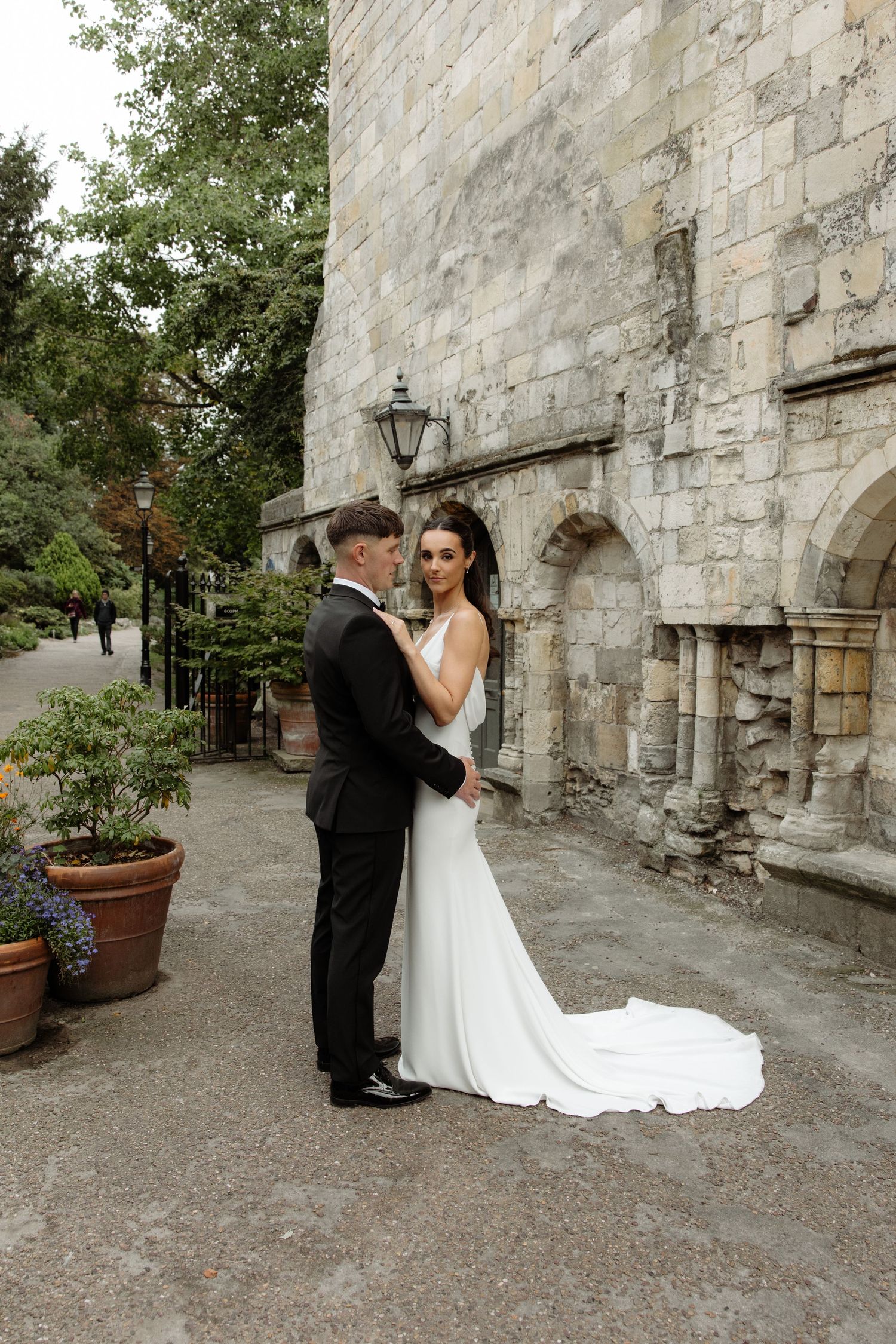 A romantic wedding couple shares an intimate moment against a rustic stone wall backdrop at a historic venue.