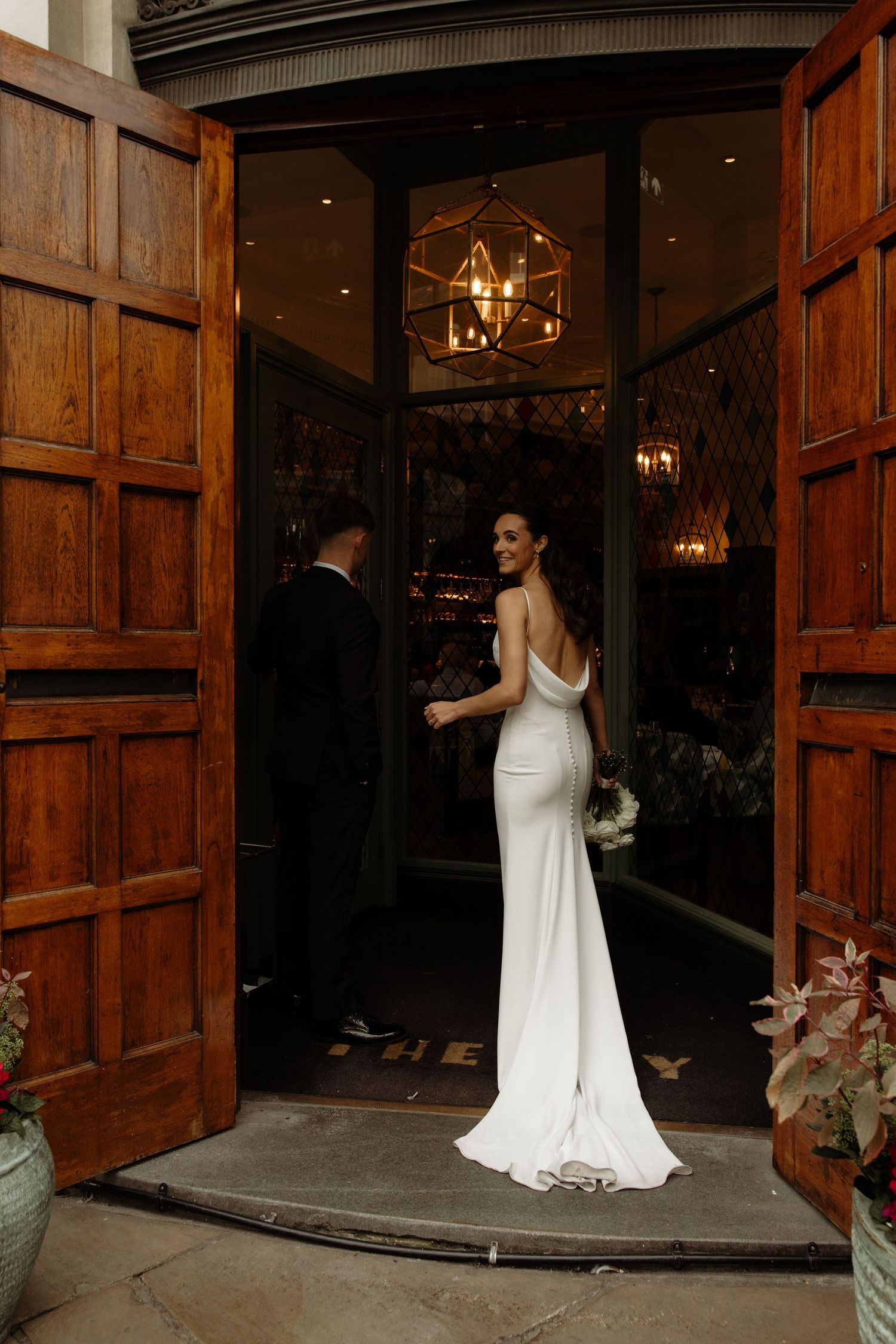 Bride in white wedding dress enters rustic wooden doors at dusk with glowing lights in background.