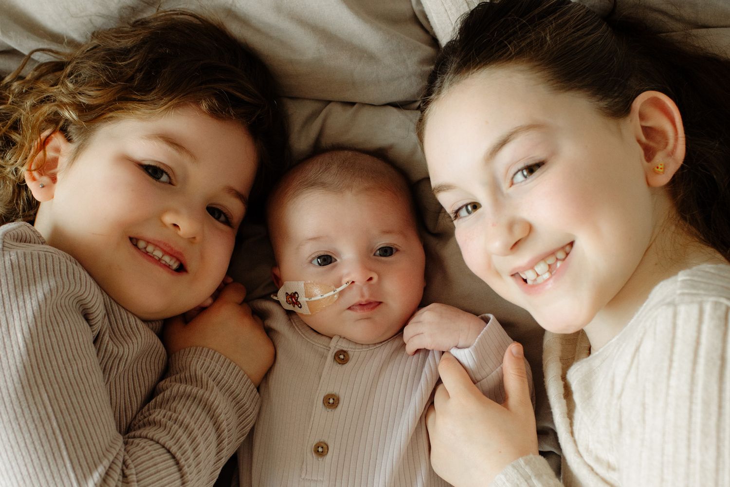 Three young siblings lying together on a bed, including a baby with a feeding tube, smiling warmly.