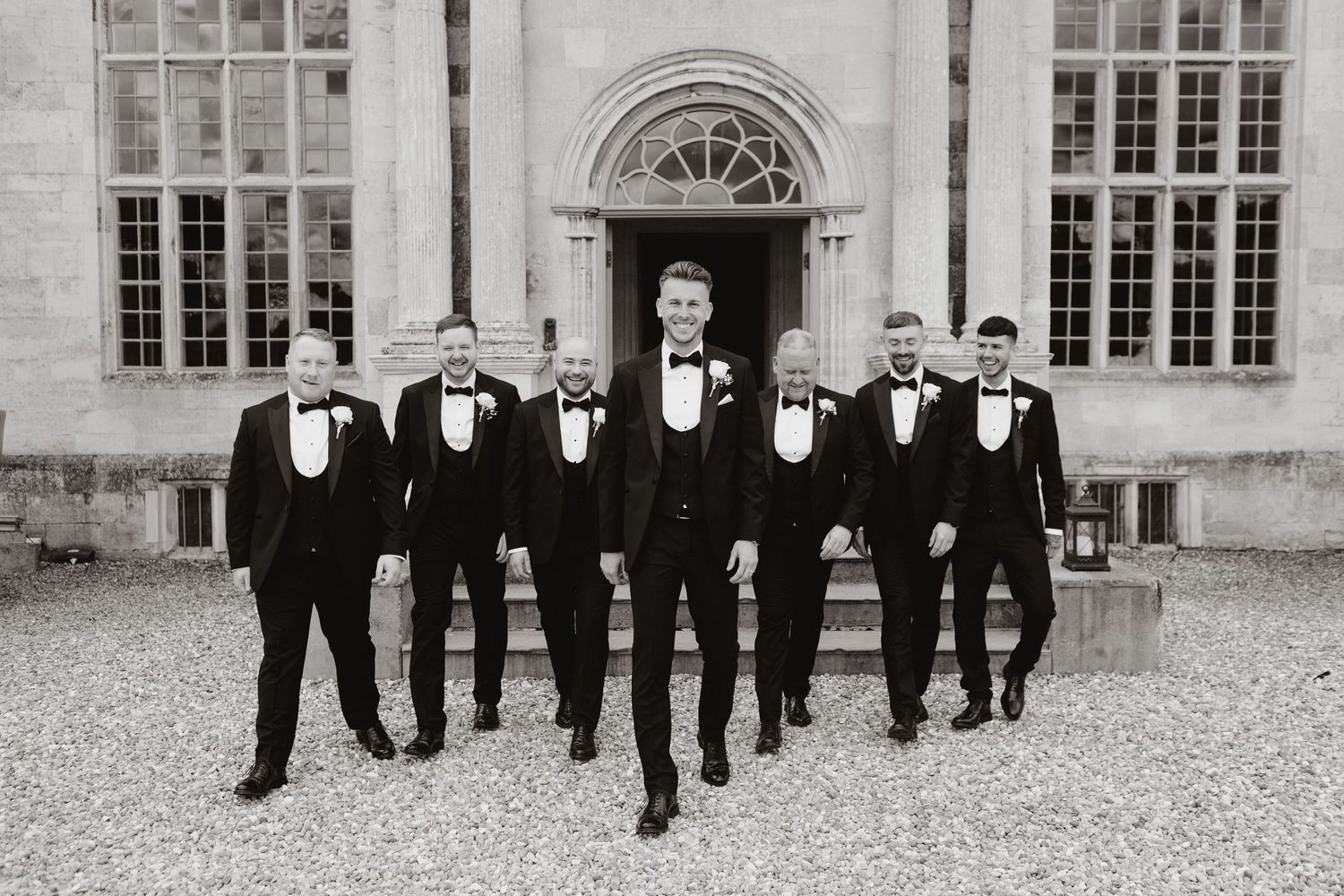 A group of elegantly dressed guests in black tuxedos walk along a paved courtyard outside a historic building.