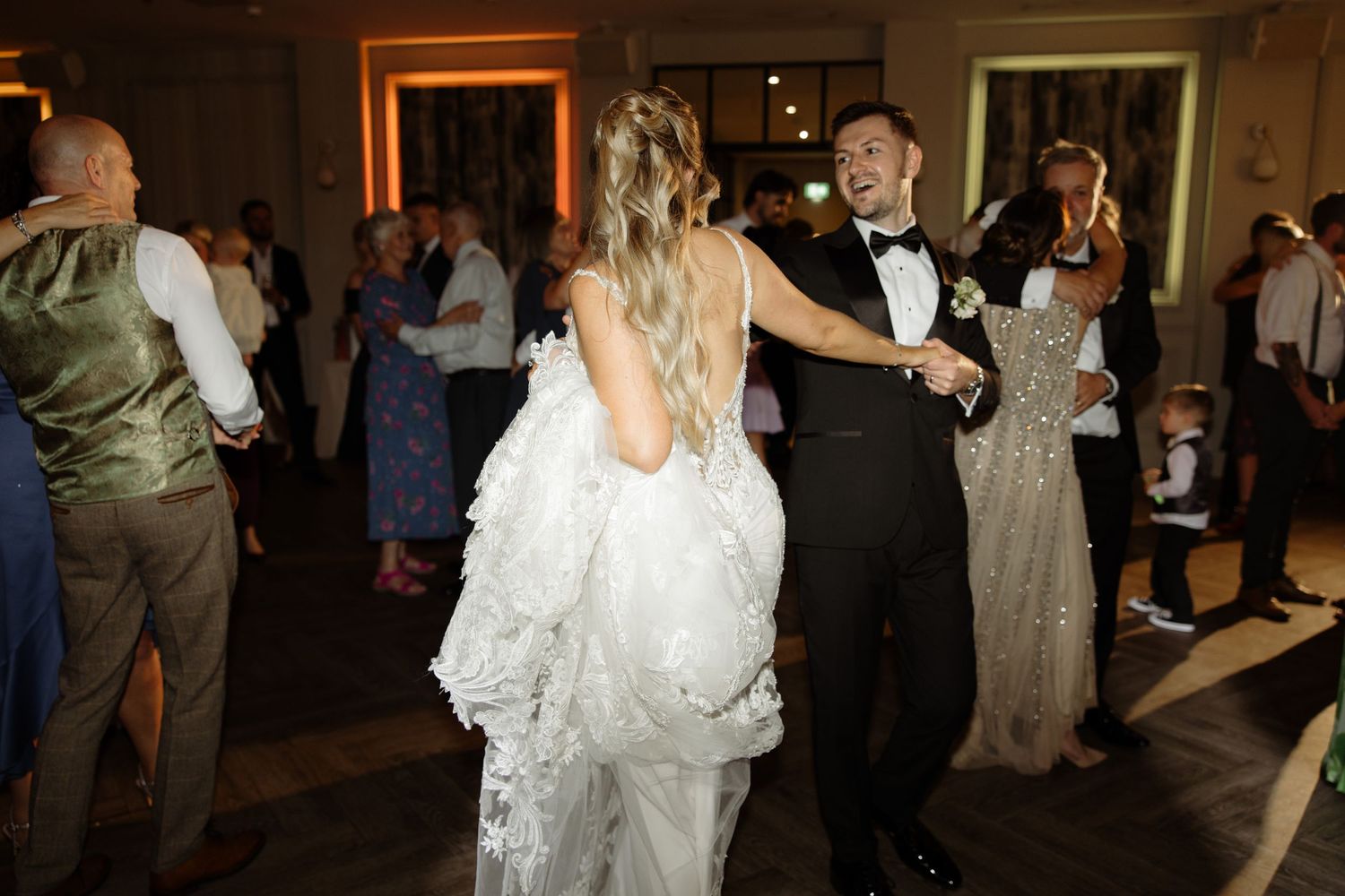 Wedding guests dance together on a dimly lit dance floor during an evening reception celebration.