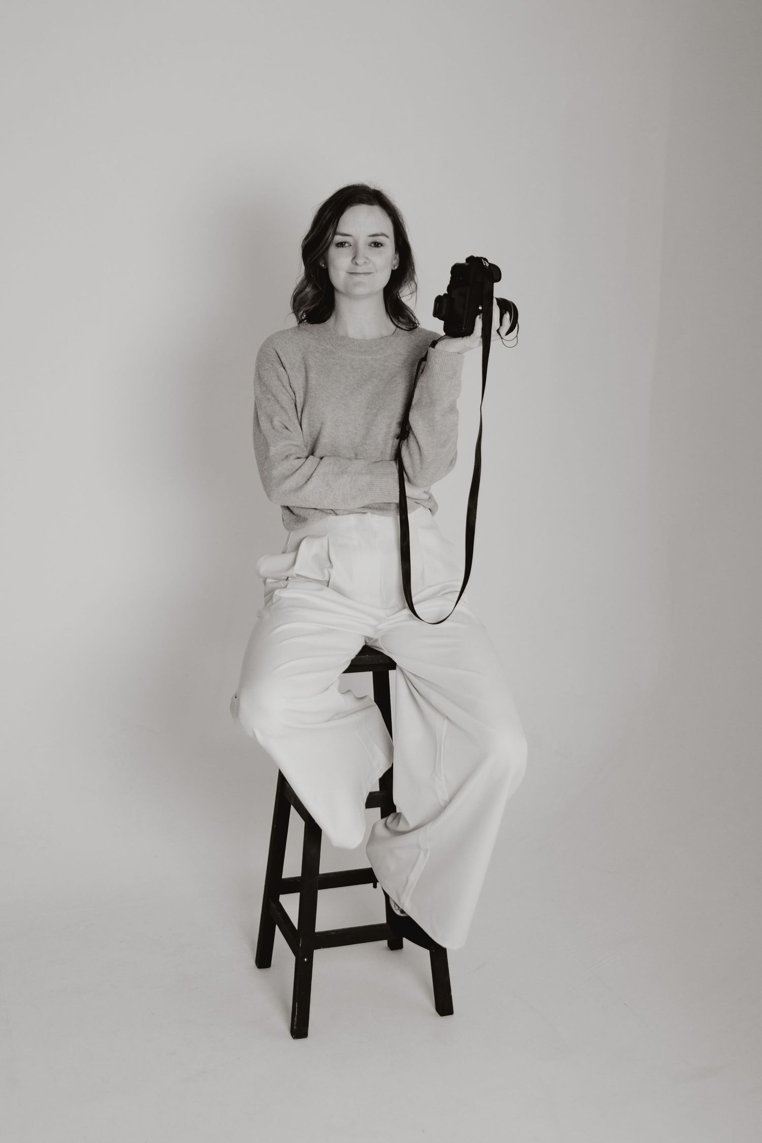 A photographer in casual attire sits on a wooden stool against a light backdrop.