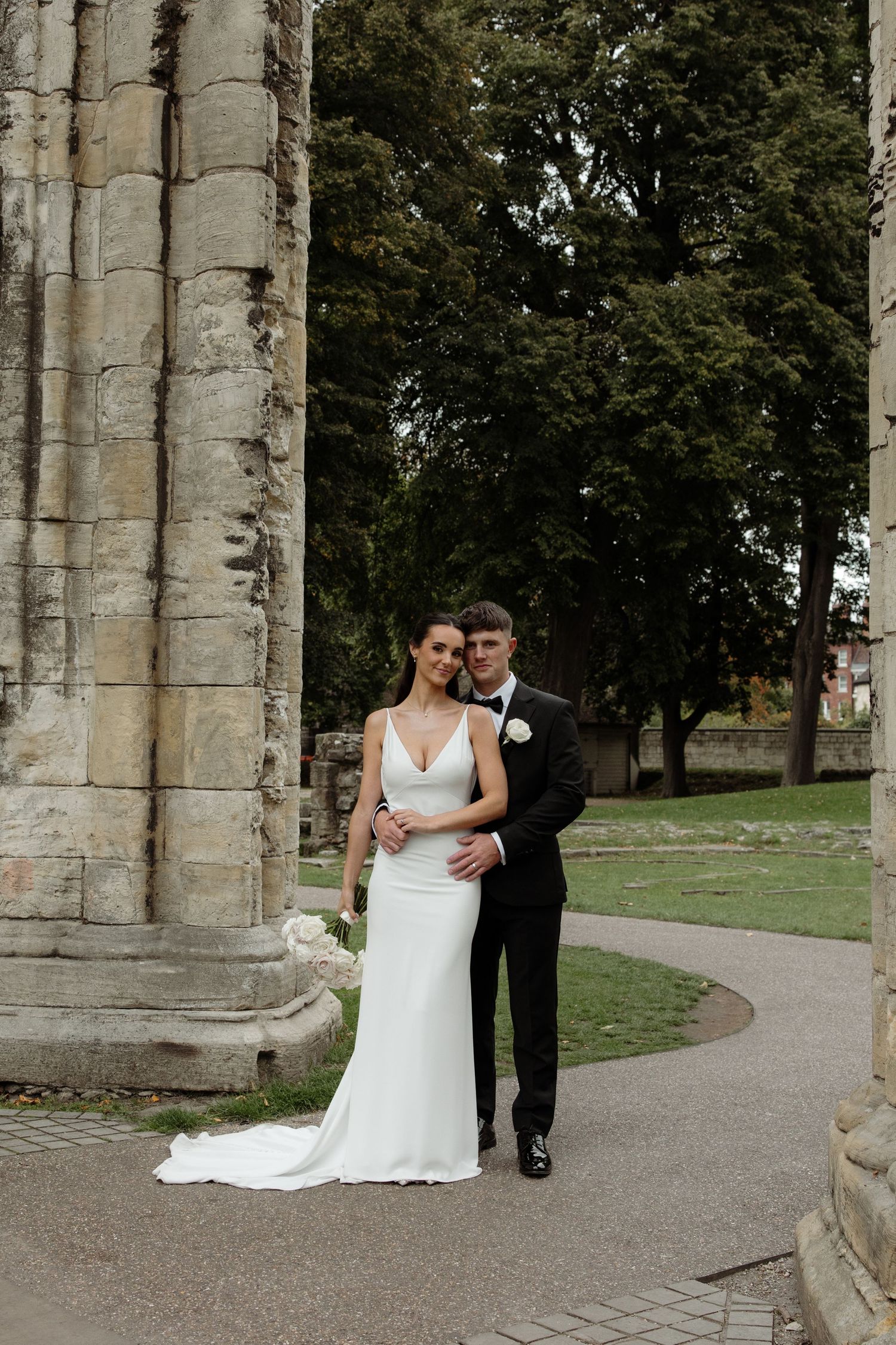 A couple in wedding attire poses between stone columns in an outdoor garden setting.