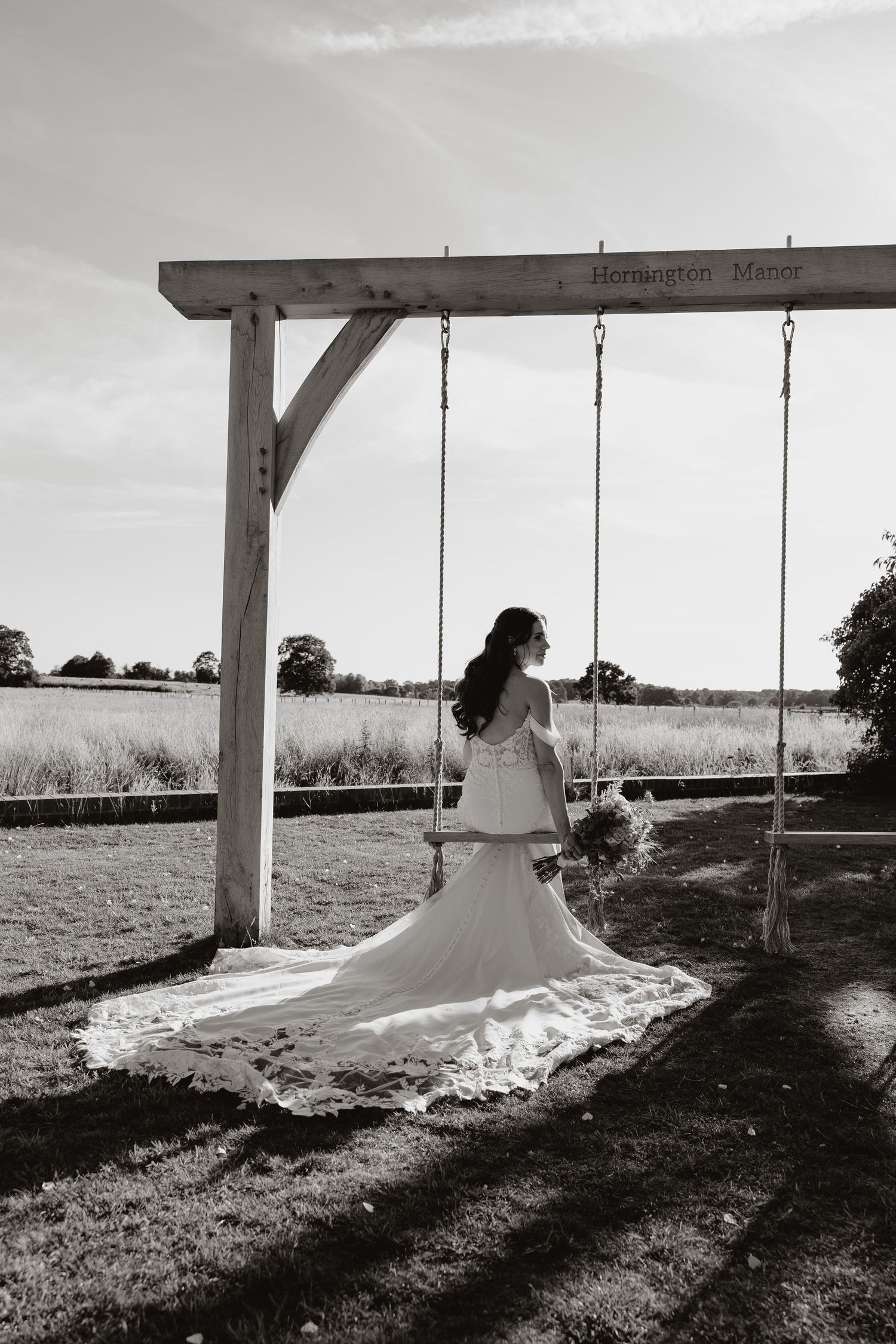A person in a flowing white wedding dress sits peacefully on a wooden swing in a rustic outdoor setting.