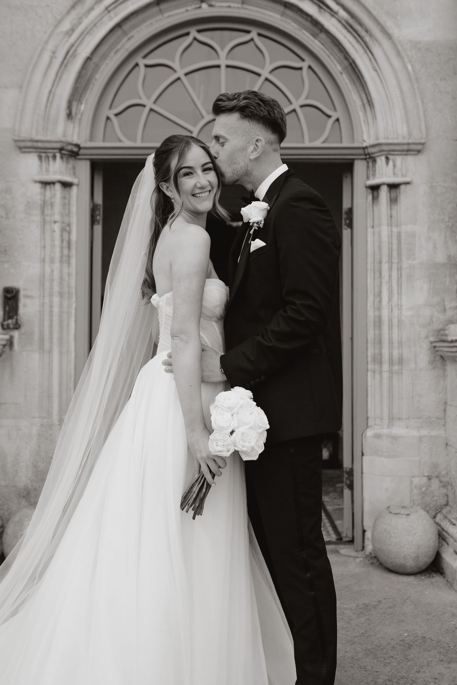 Black and white wedding portrait of newlyweds standing in front of an ornate arched church doorway.