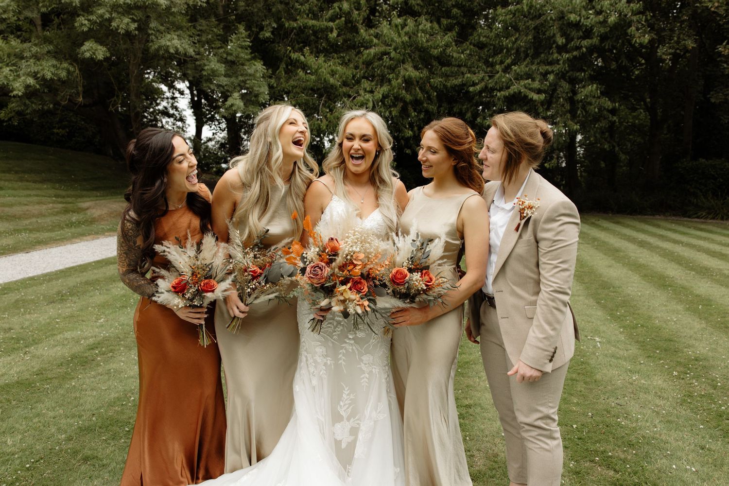 A wedding party stands on a lush green lawn sharing candid moments and laughter while dressed in earth-toned attire.