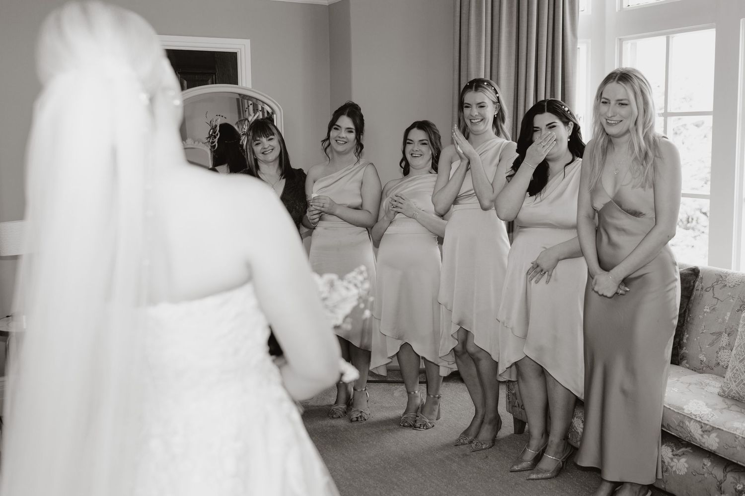 A bride in a white wedding dress stands surrounded by bridesmaids in blush pink dresses in a bright sunlit room.