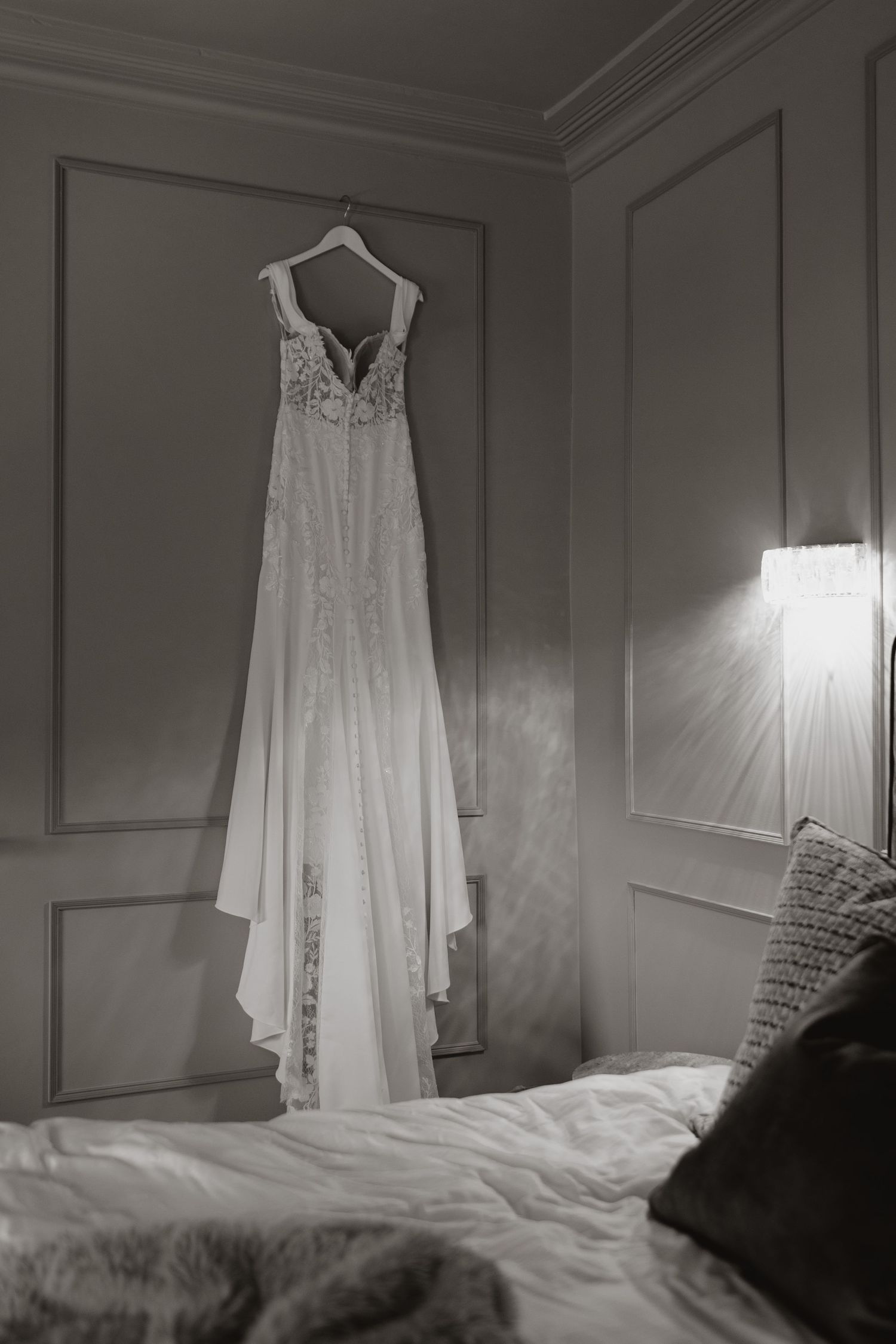 A delicate white lace wedding dress hangs elegantly on a hanger against a soft gray wall near a bed in natural light.