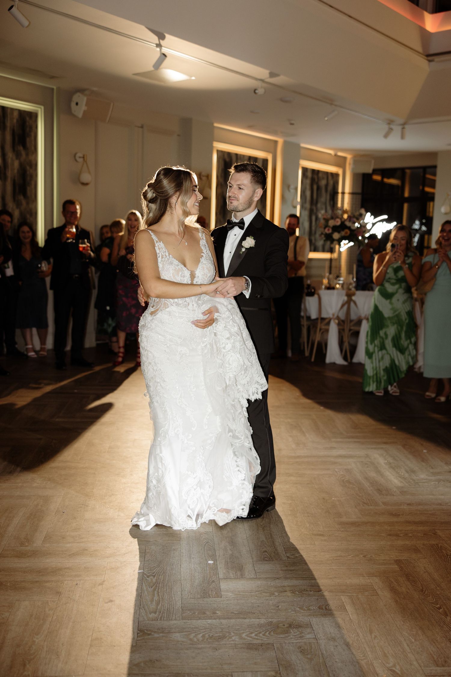 A couple performs their first dance at a wedding reception on a wooden dance floor surrounded by guests.