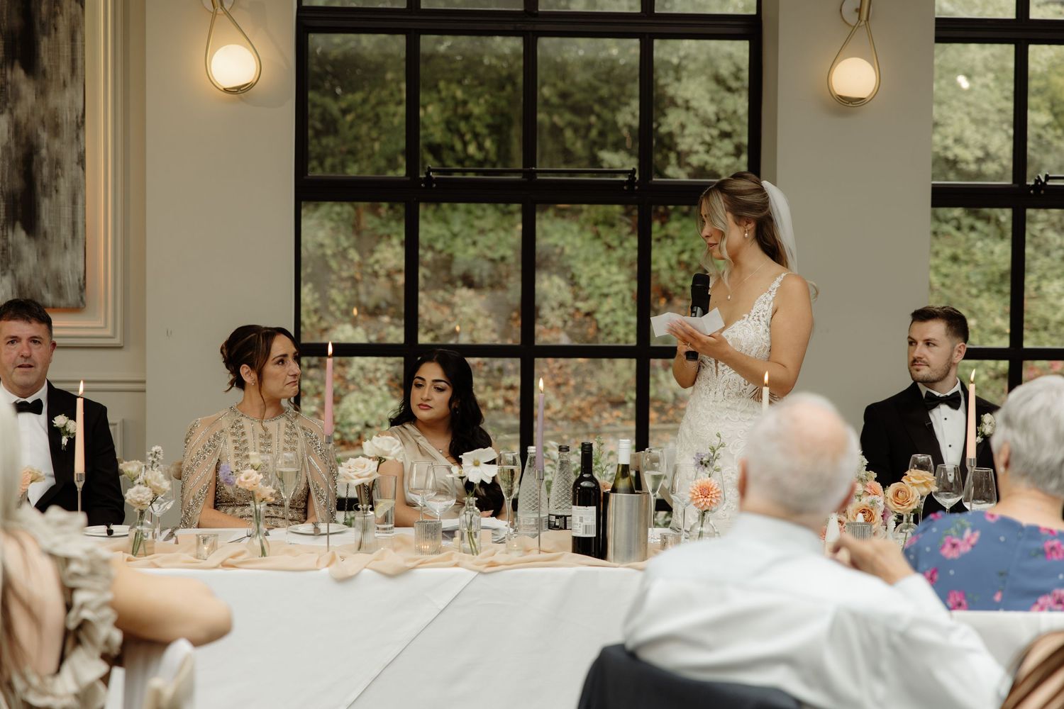 A wedding reception speech being given at an elegant indoor venue with large windows and pendant lighting.