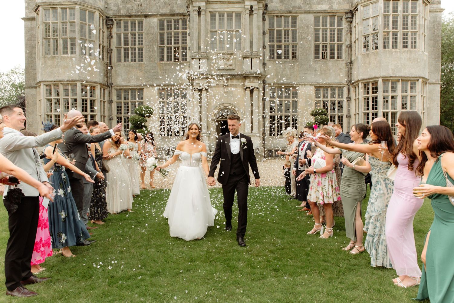 Wedding guests toss white flower petals in celebration outside a grand stone manor building during a summer ceremony.