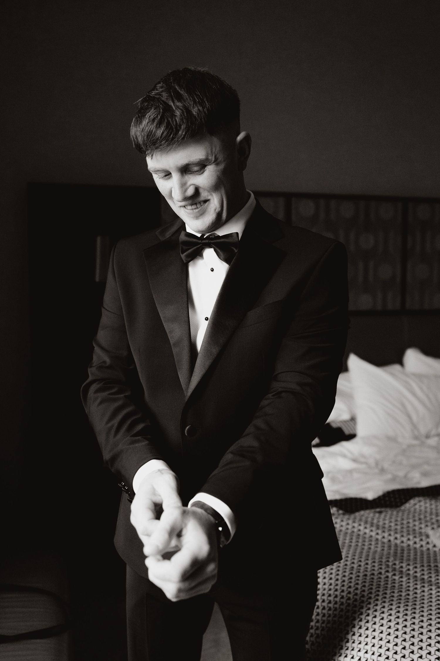 A person in a black suit adjusts their cuffs while getting dressed in a dimly lit room.