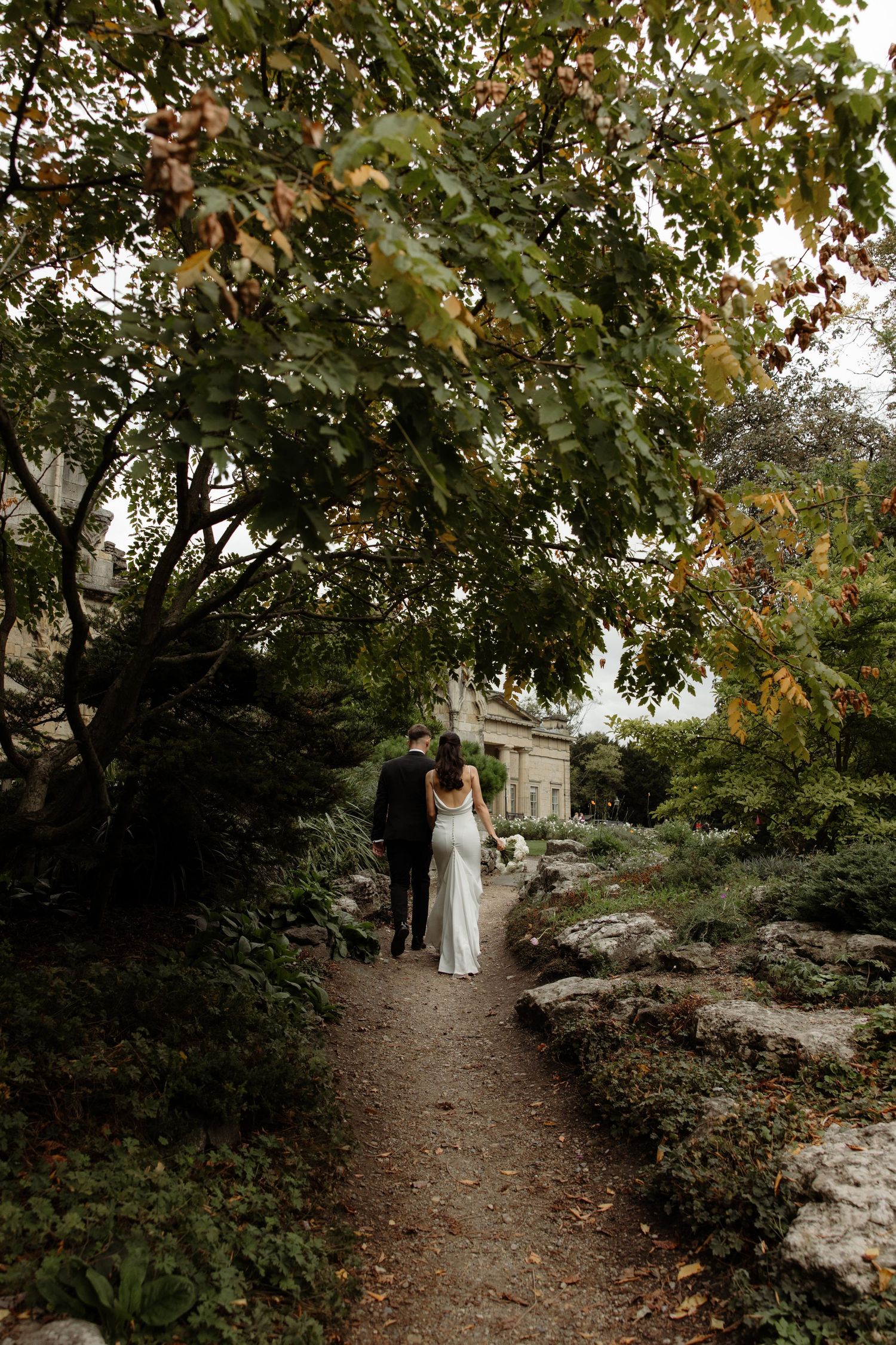 A couple walks along a winding dirt path through autumn trees and foliage in a romantic outdoor setting.