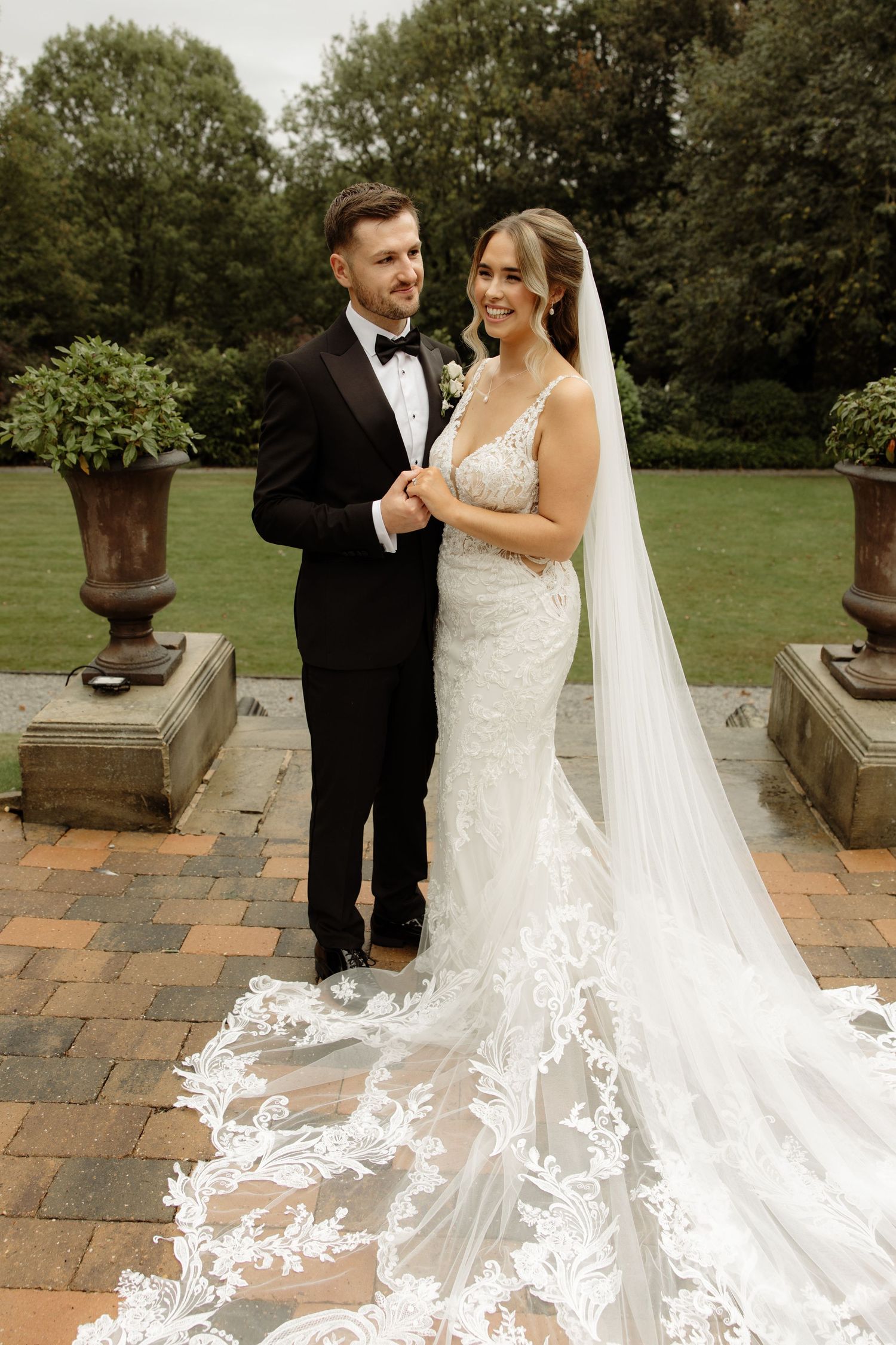 A romantic wedding couple poses on stone terrace with a flowing lace veil trailing behind them in an elegant garden setting.