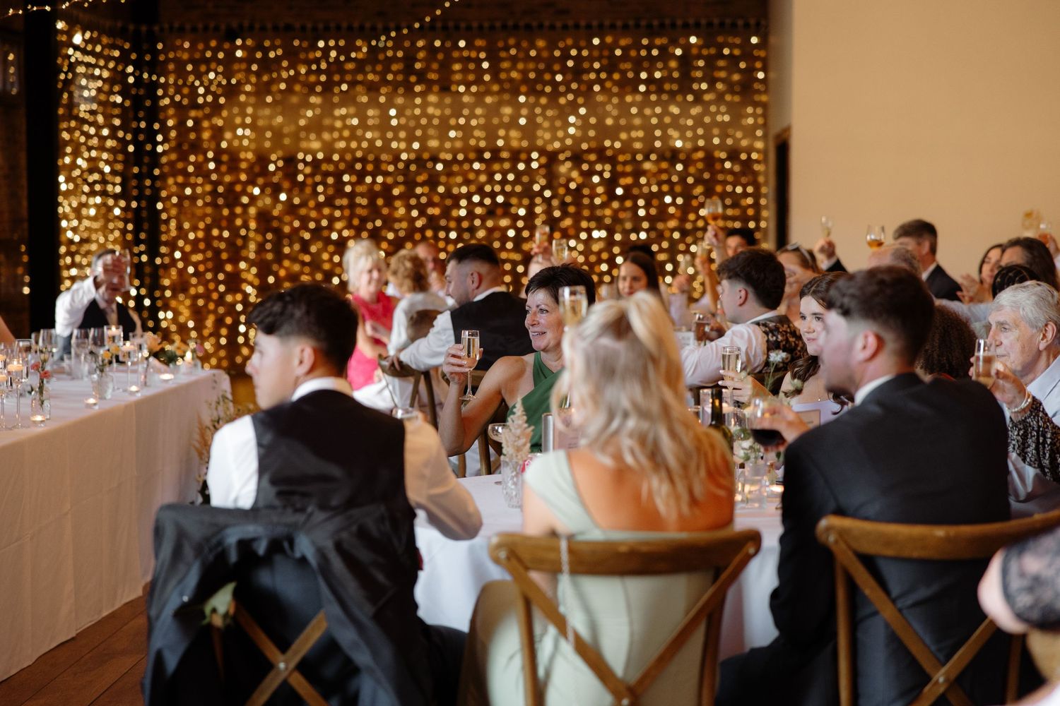 Warm string lights create a romantic backdrop at an indoor wedding reception with guests seated at tables.