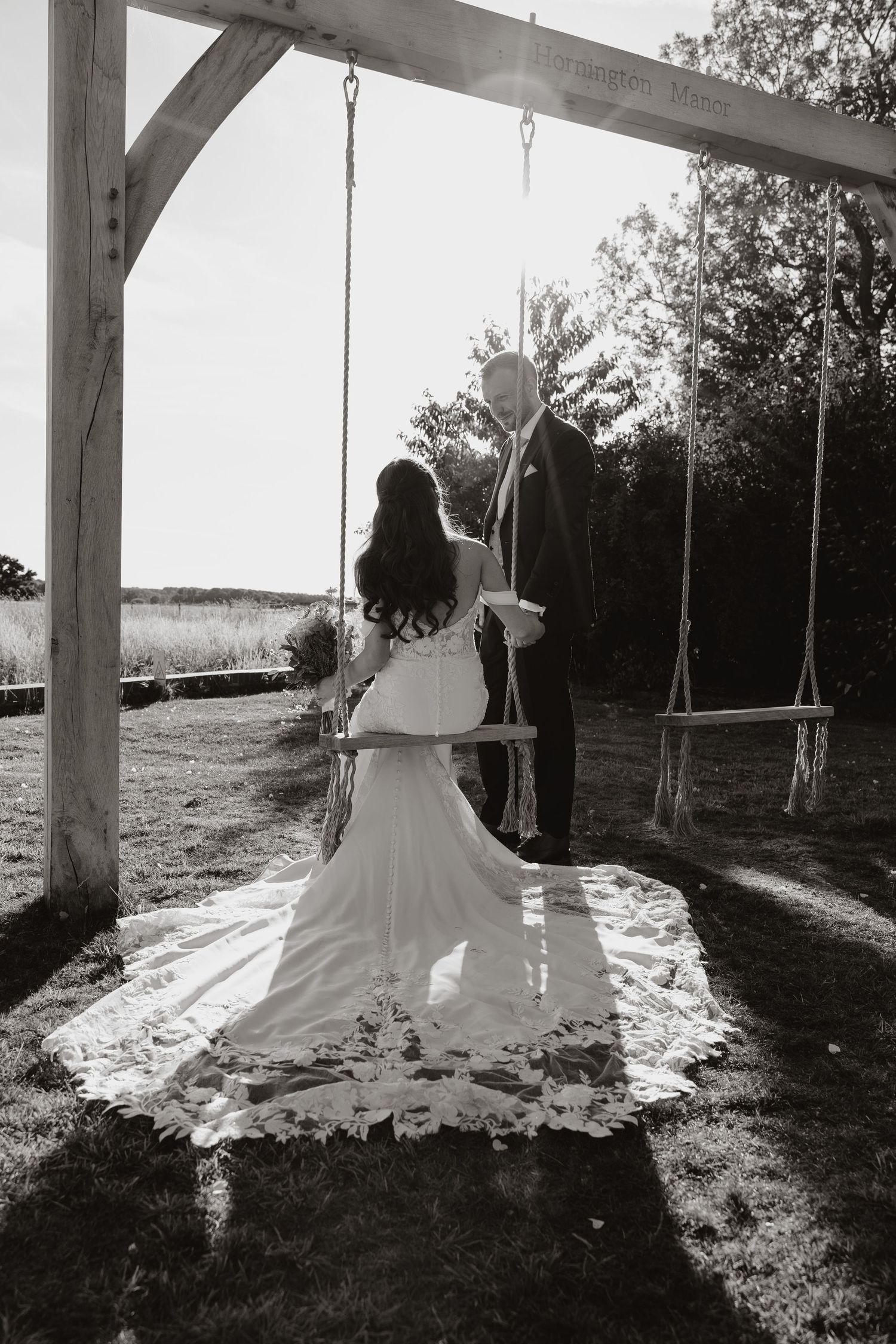 Silhouette of a person in a flowing dress sitting on a swing against sunlit trees in a black and white photo series.