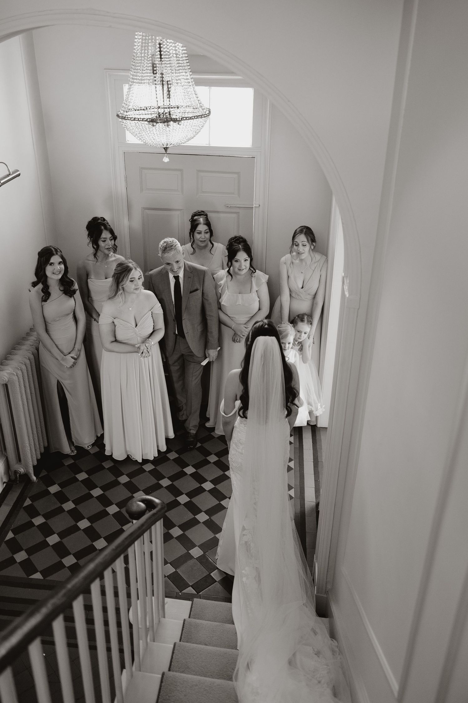 Black and white wedding photograph showing a bride in a veil on stairs with bridesmaids in matching gowns looking on.